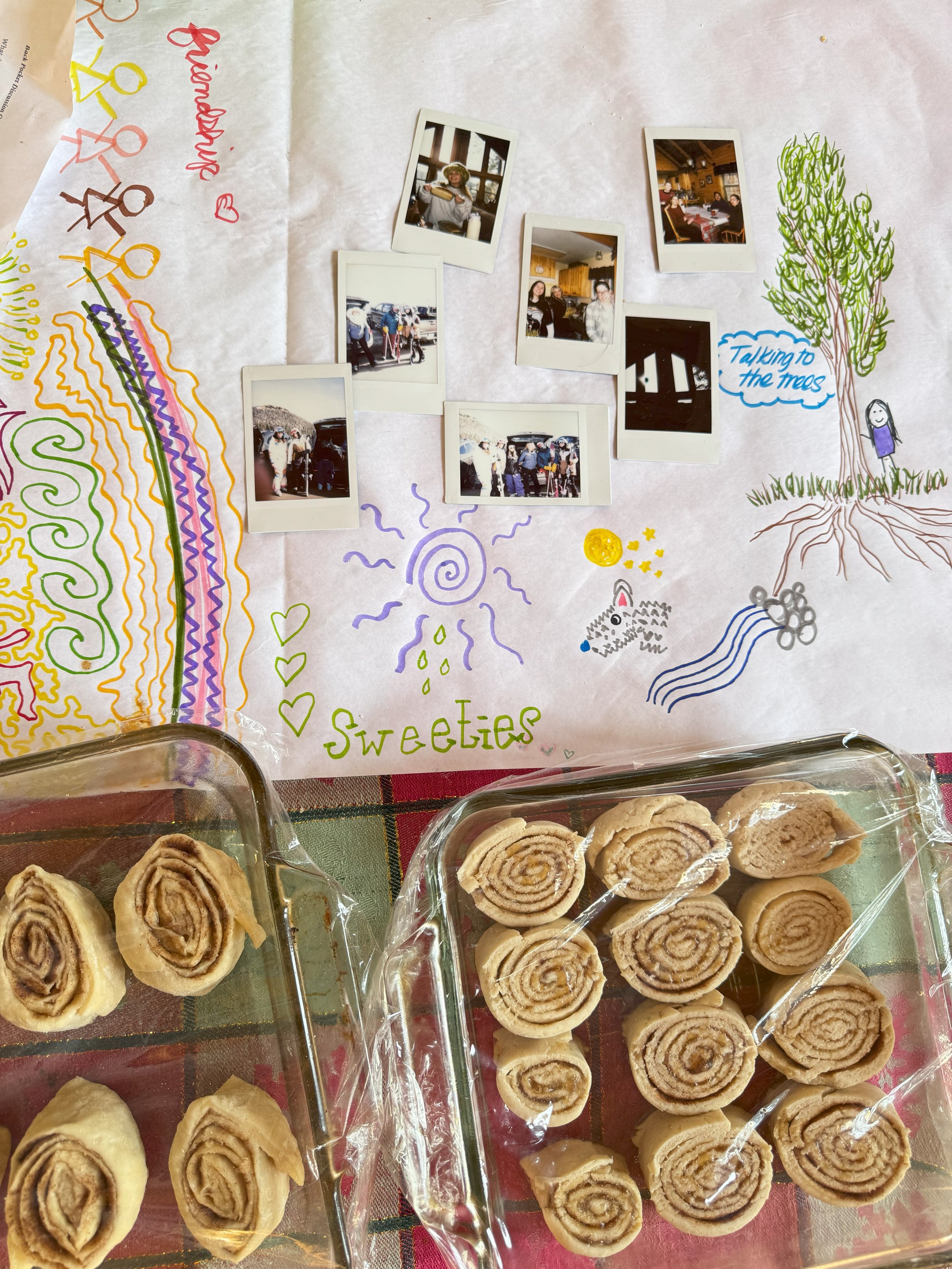 Colorful drawing with trees, rainbows, people, and words like 'Sweeties' and 'talking to the trees' on a large sheet of paper, with two trays of cinnamon rolls at the bottom.