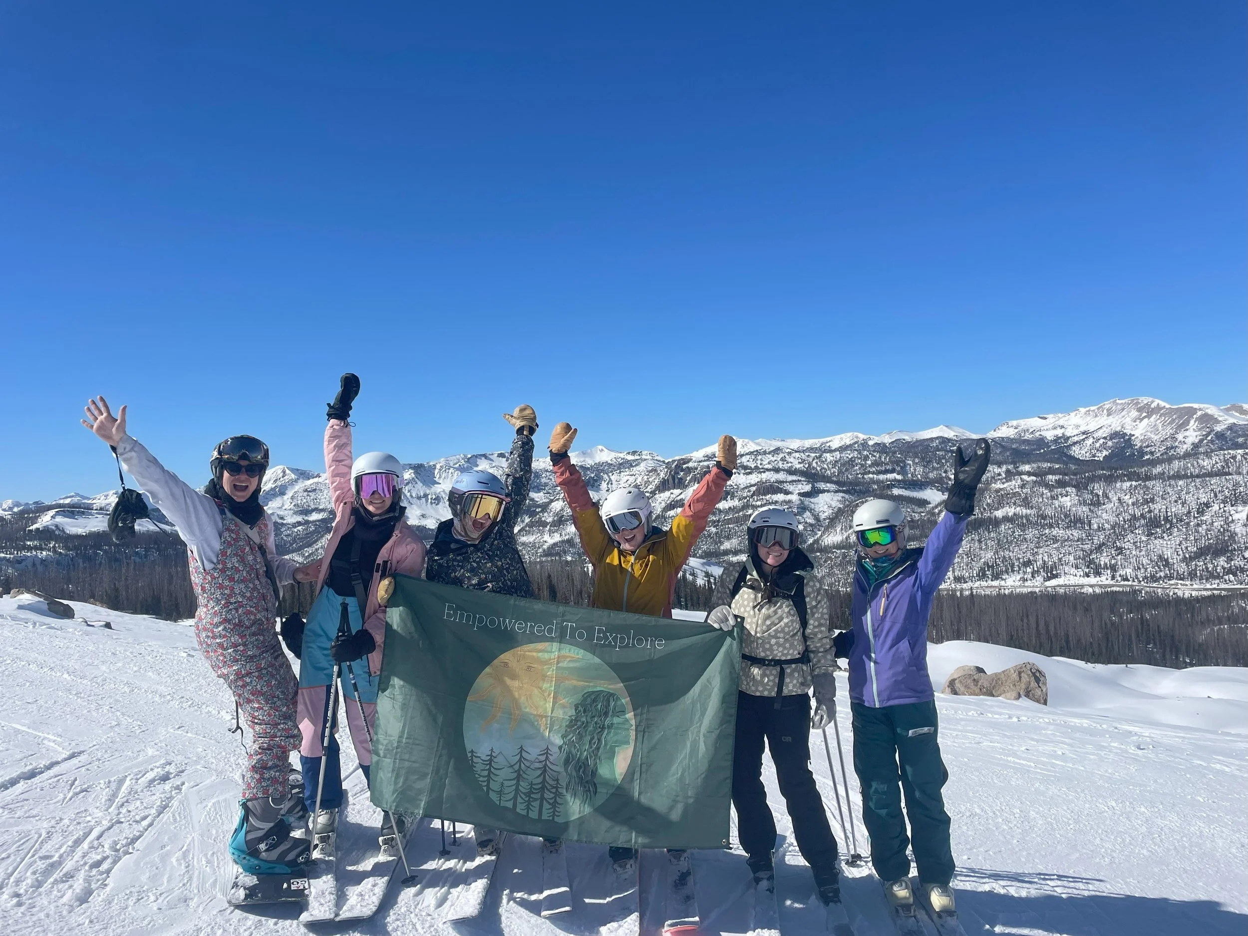 Six people in ski gear standing on snow on a mountain, holding a banner that reads "Empowered To Explore." They are celebrating with their arms raised, with snow-covered mountains in the background under a clear blue sky.