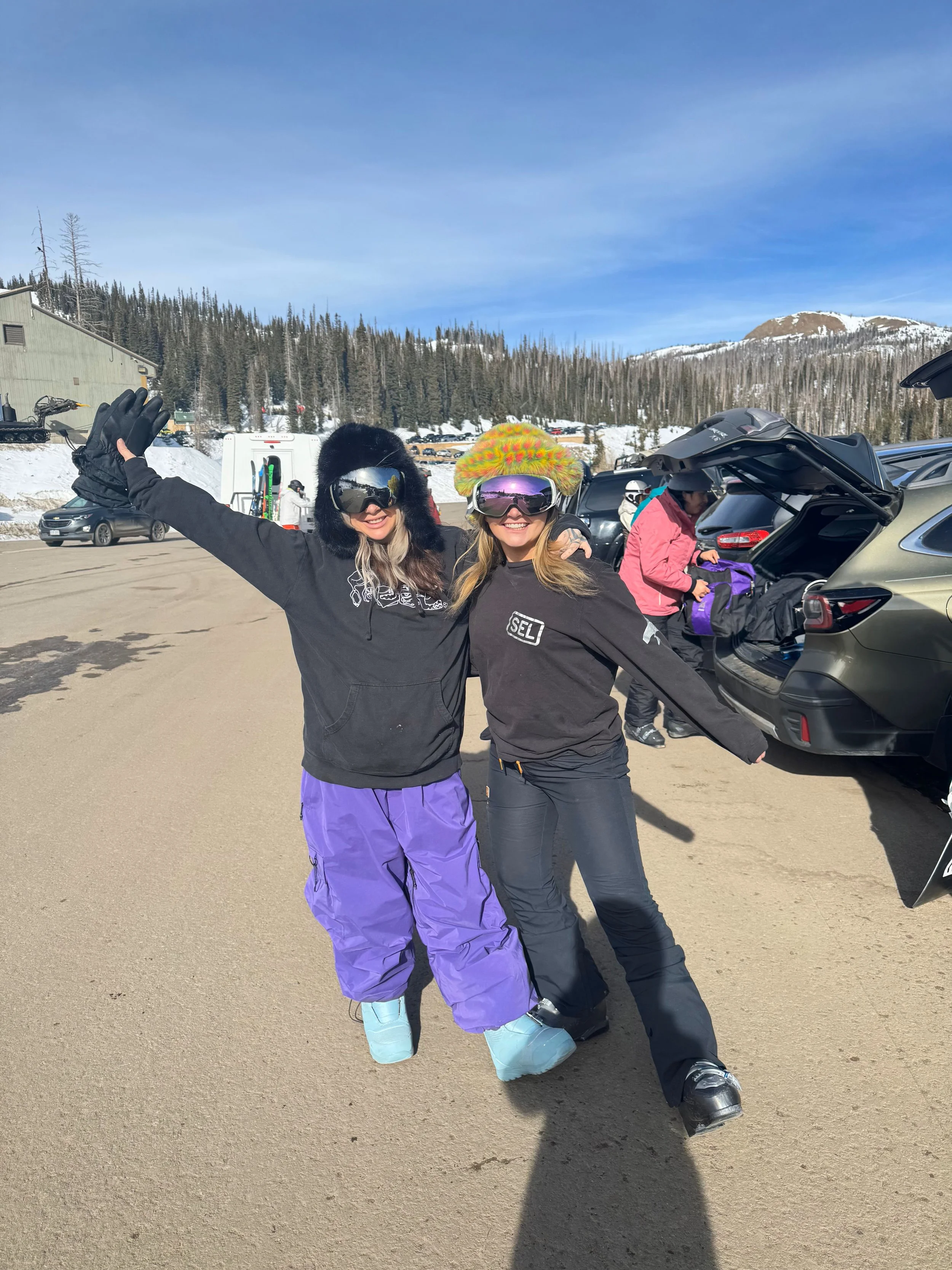 Two women in winter skiing gear smiling and posing for the camera in a snowy mountain parking lot.
