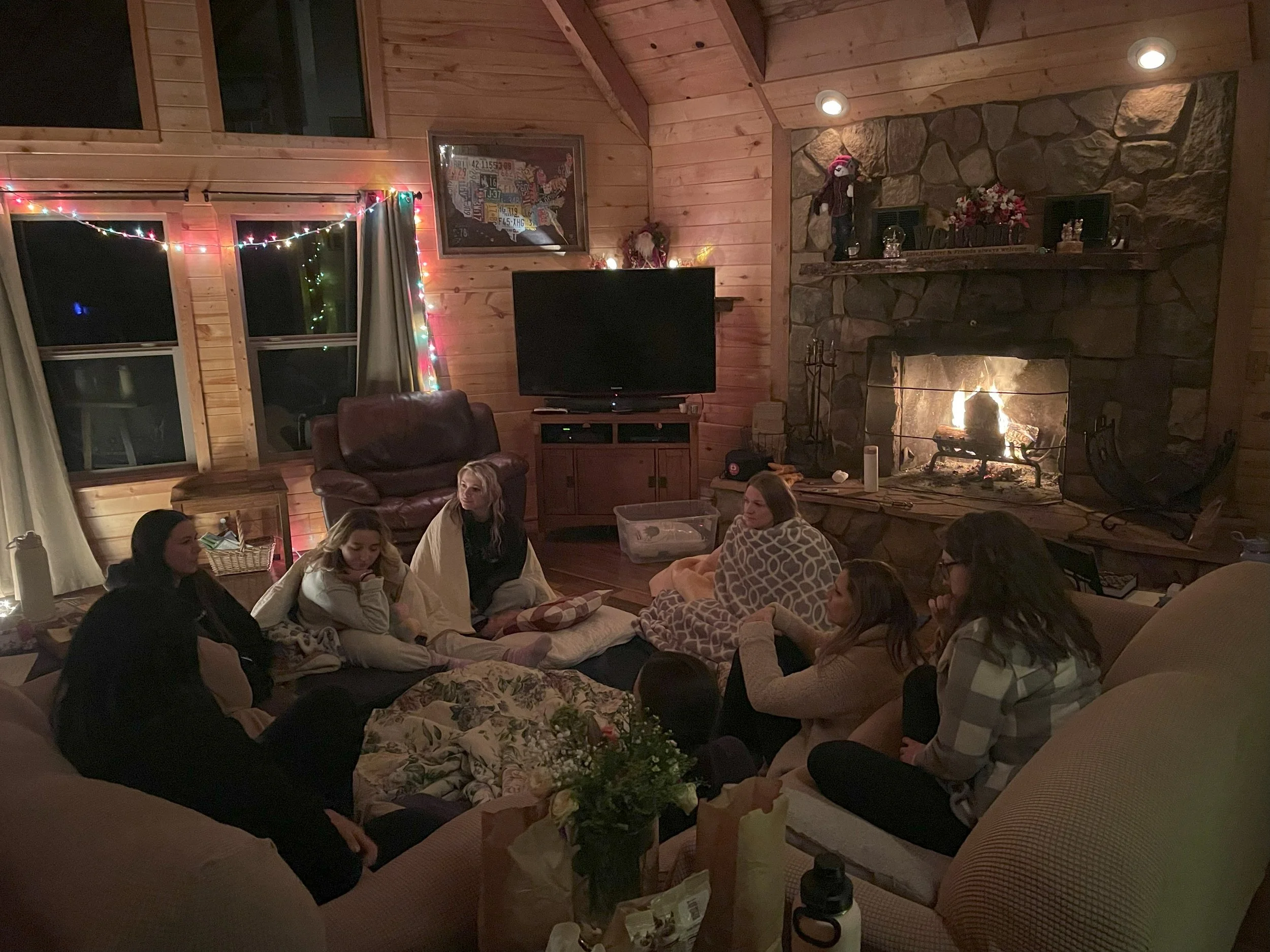 A group of people gathered in a cozy living room with wooden walls, sitting on a large sectional sofa near a fireplace with a fire burning. There are string lights hanging around the windows, creating a warm atmosphere. The room has a television on a stand, a leather armchair, and various decorations including framed art and flowers.