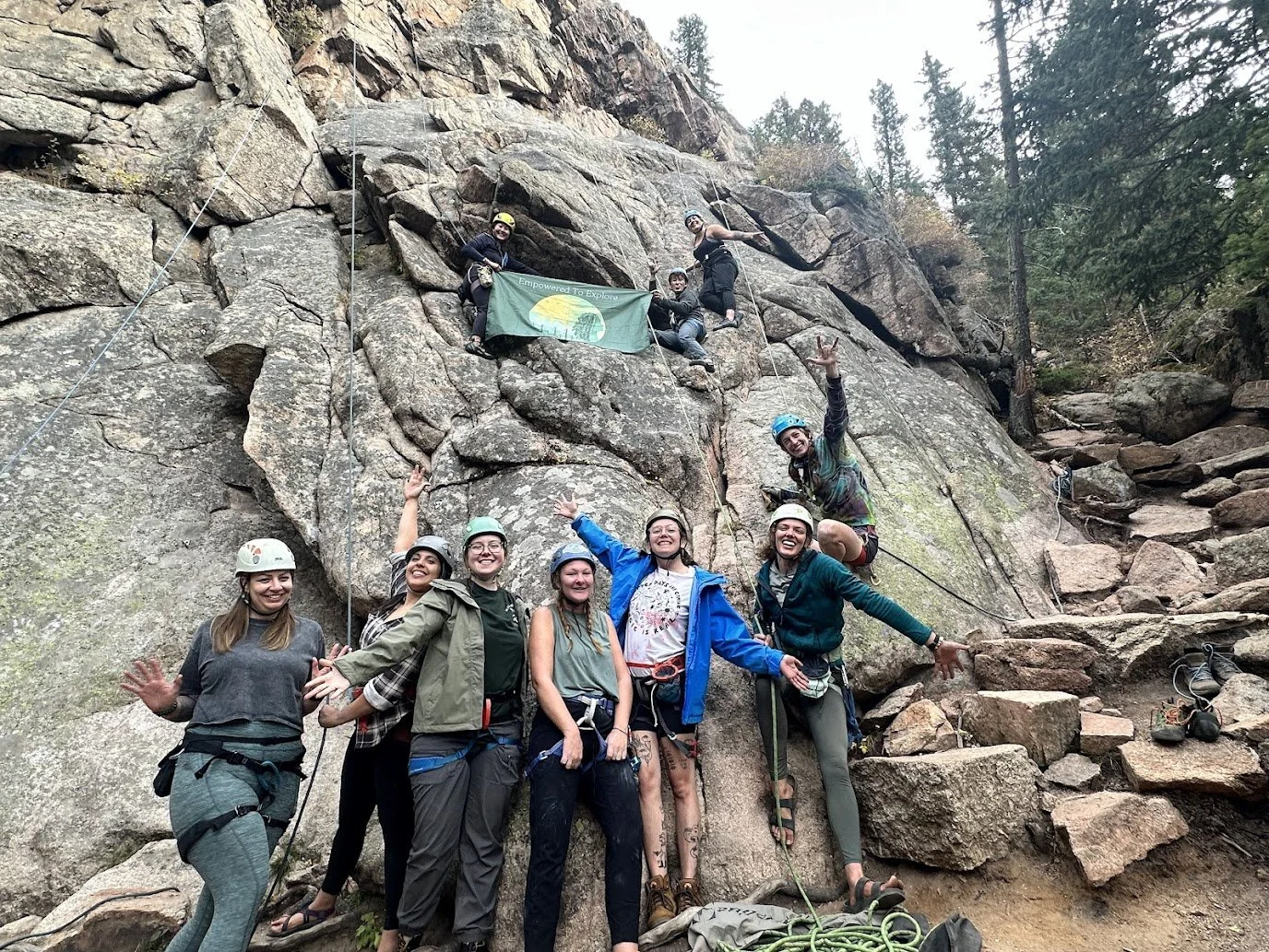 Group of seven outdoor rock climbers with helmets and harnesses smiling and celebrating at the base of a rock face in a forested area.