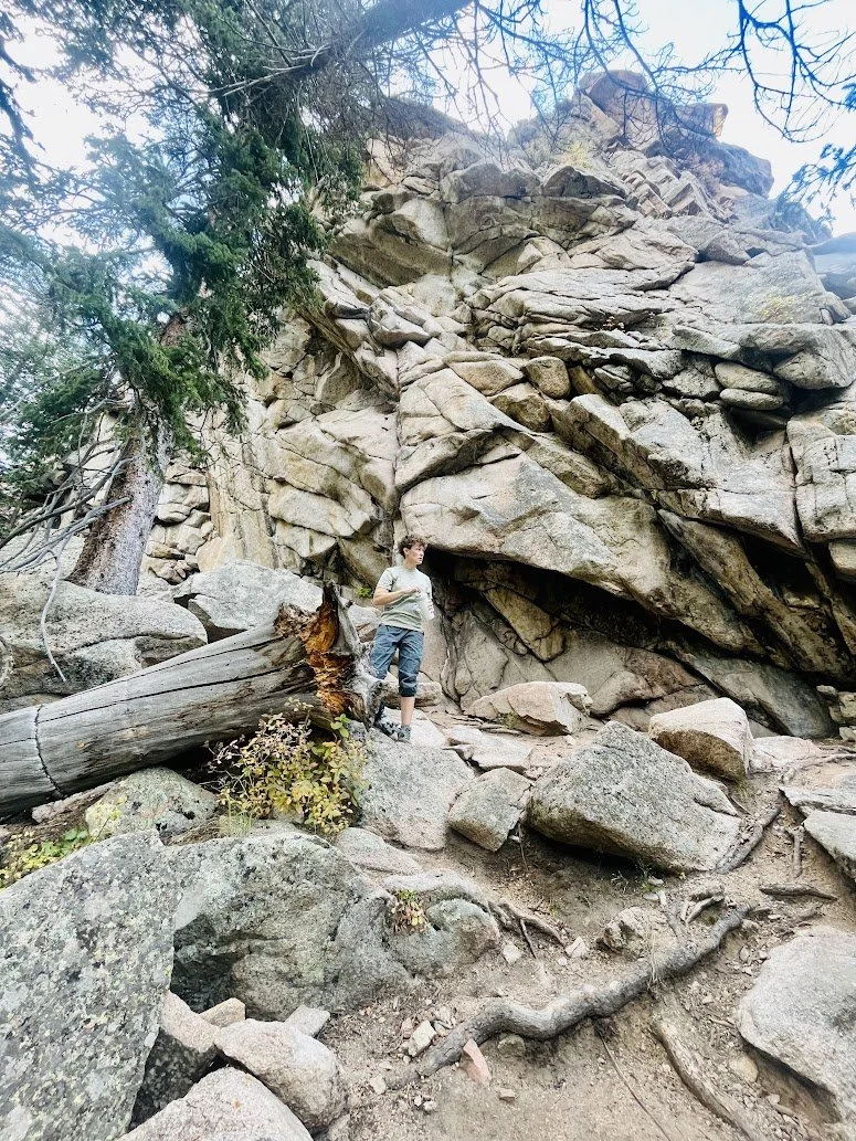 A young boy stands on a rocky trail in front of large boulders and a rocky cliff face, with trees and branches visible in the background.