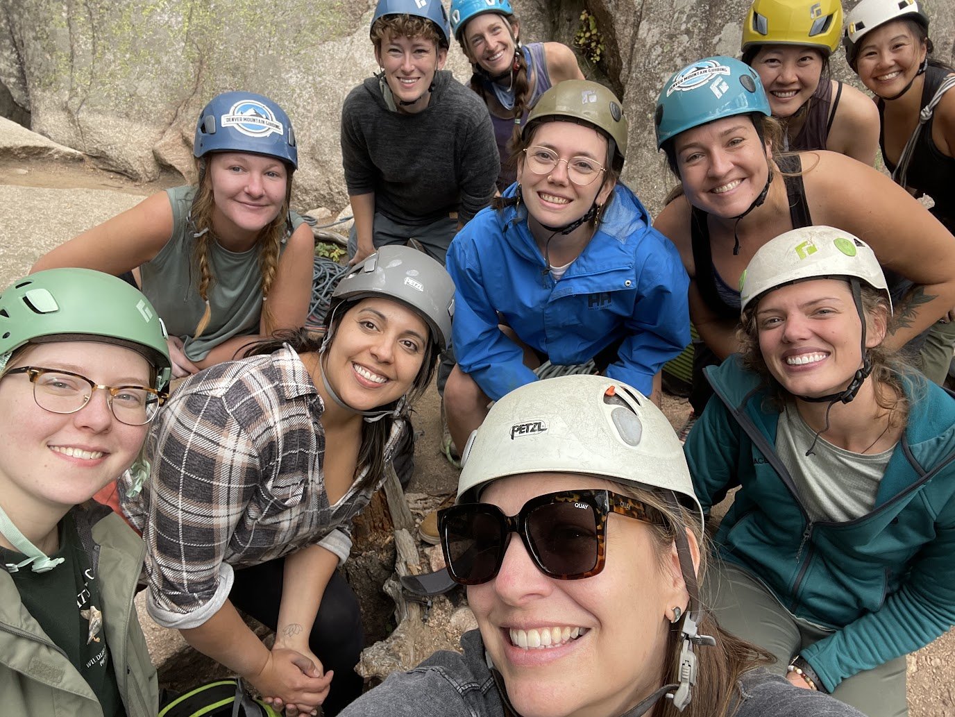 A group of women in outdoor gear and climbing helmets smiling in a rocky outdoor setting.