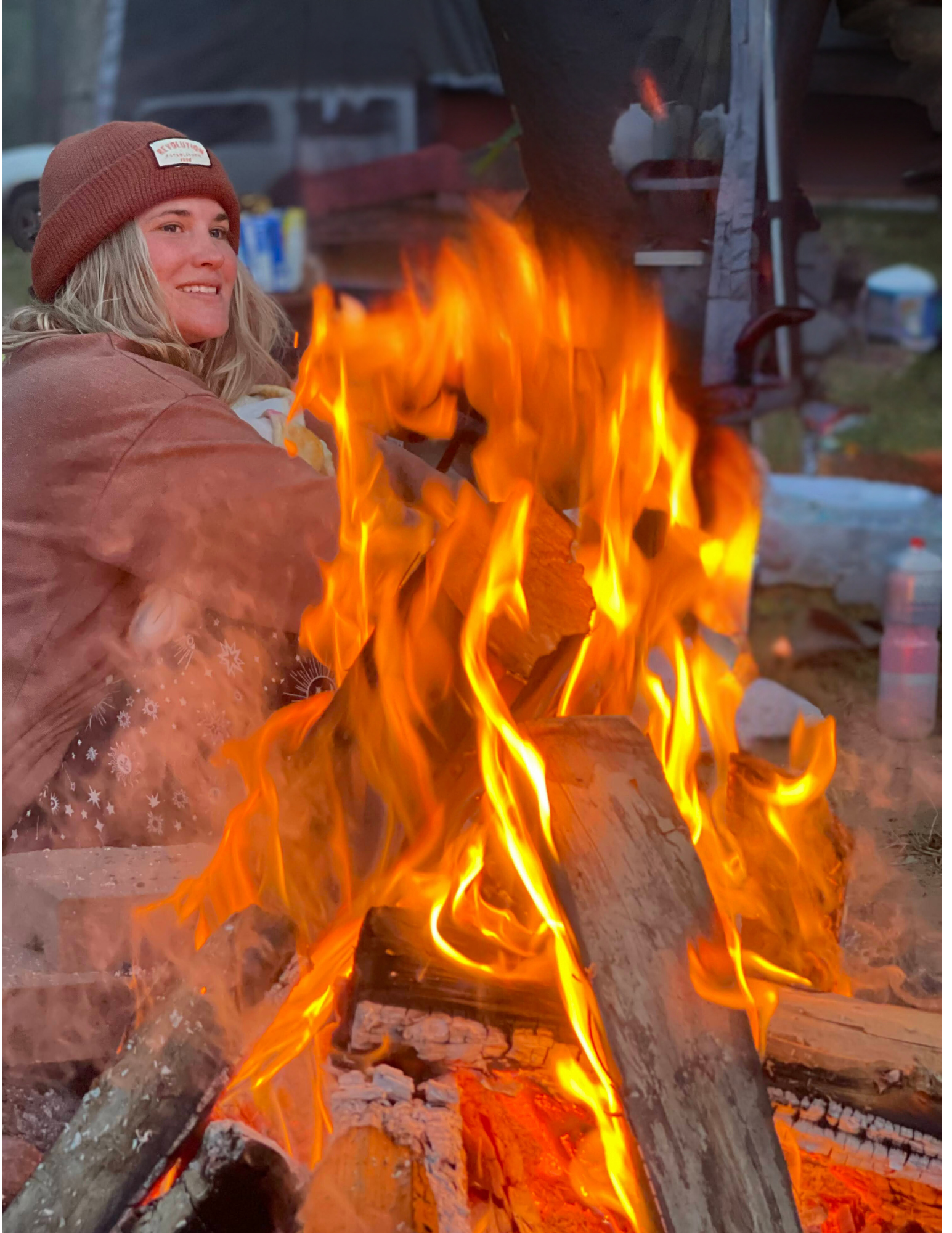 A woman in a brown beanie hat and pink jacket is sitting next to a campfire with bright orange and yellow flames, outdoors in a wooded area.