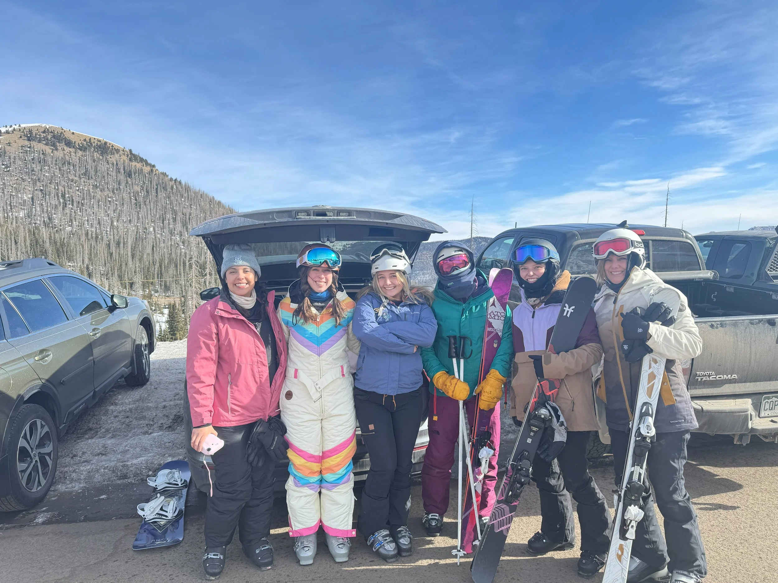 Group of six women dressed in ski gear standing outdoors in a parking lot with mountains in the background.