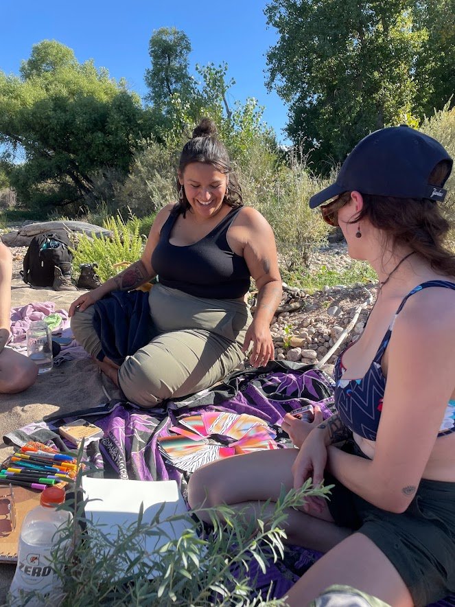 Two women sitting outdoors on a colorful blanket with art supplies, surrounded by trees and bushes, enjoying a sunny day.