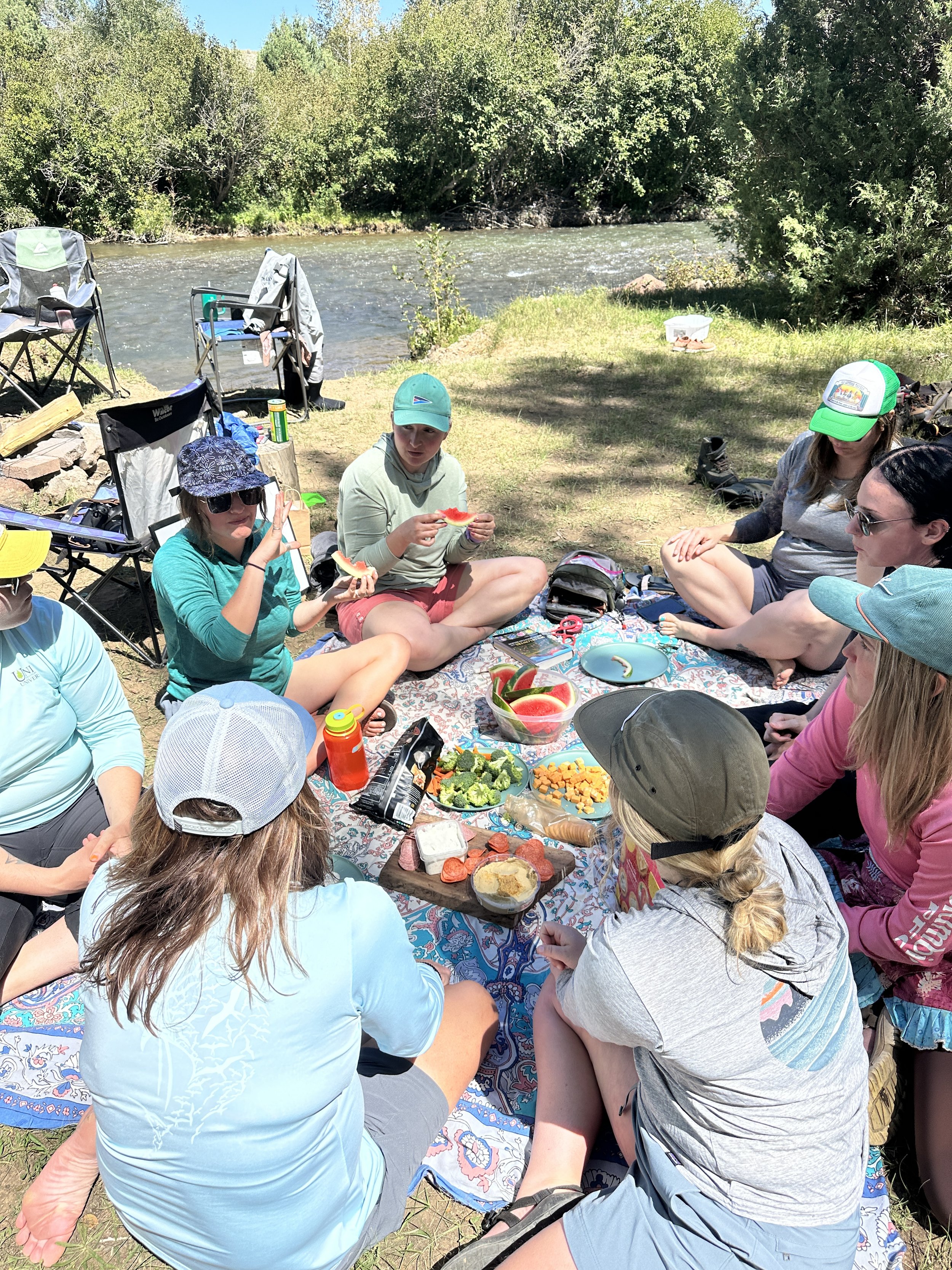 Group of people having a picnic by a river, seated on blankets with food and drinks, surrounded by trees and a sunny sky.