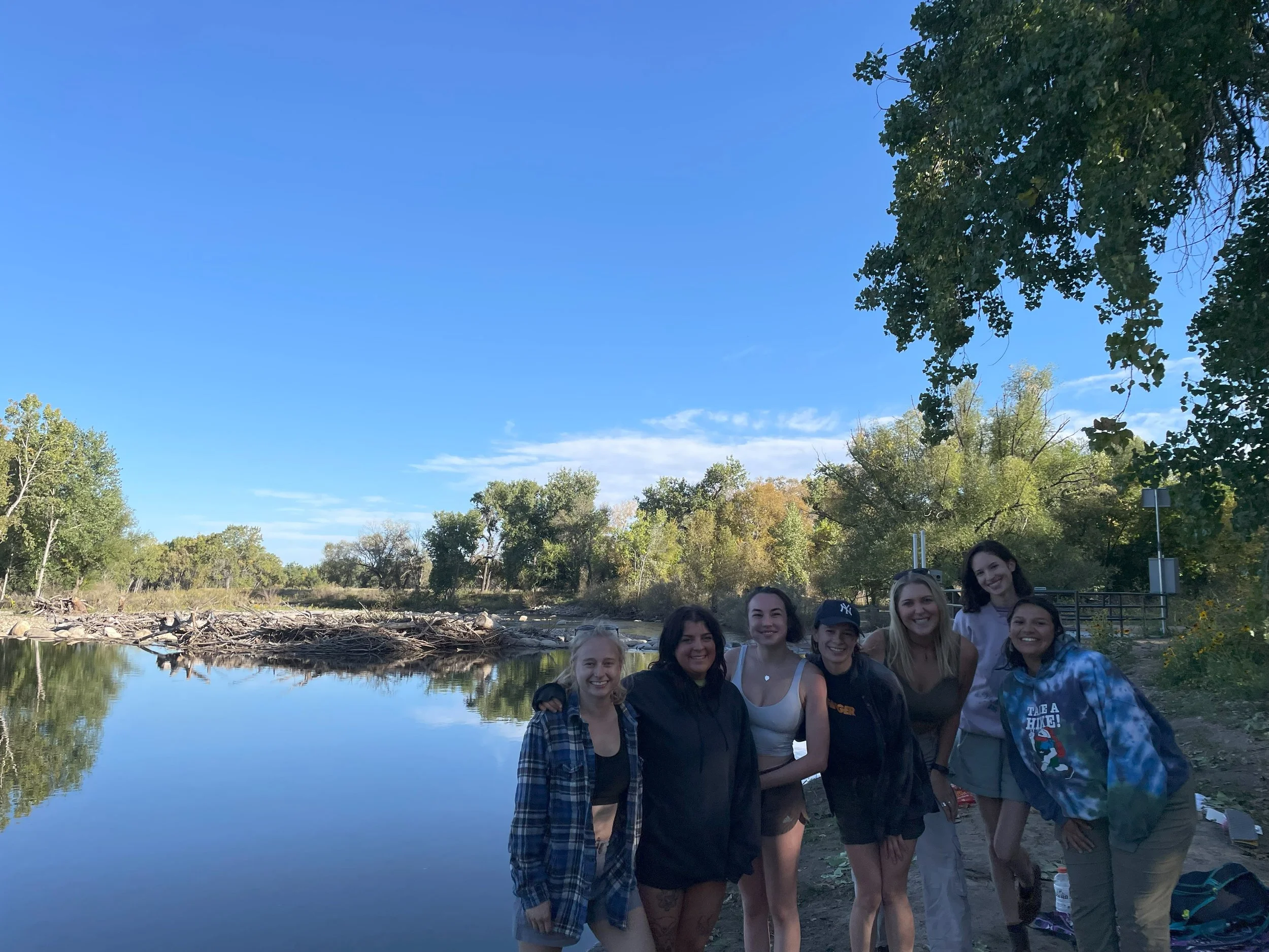 Group of seven women smiling by a river on a clear day with trees in the background