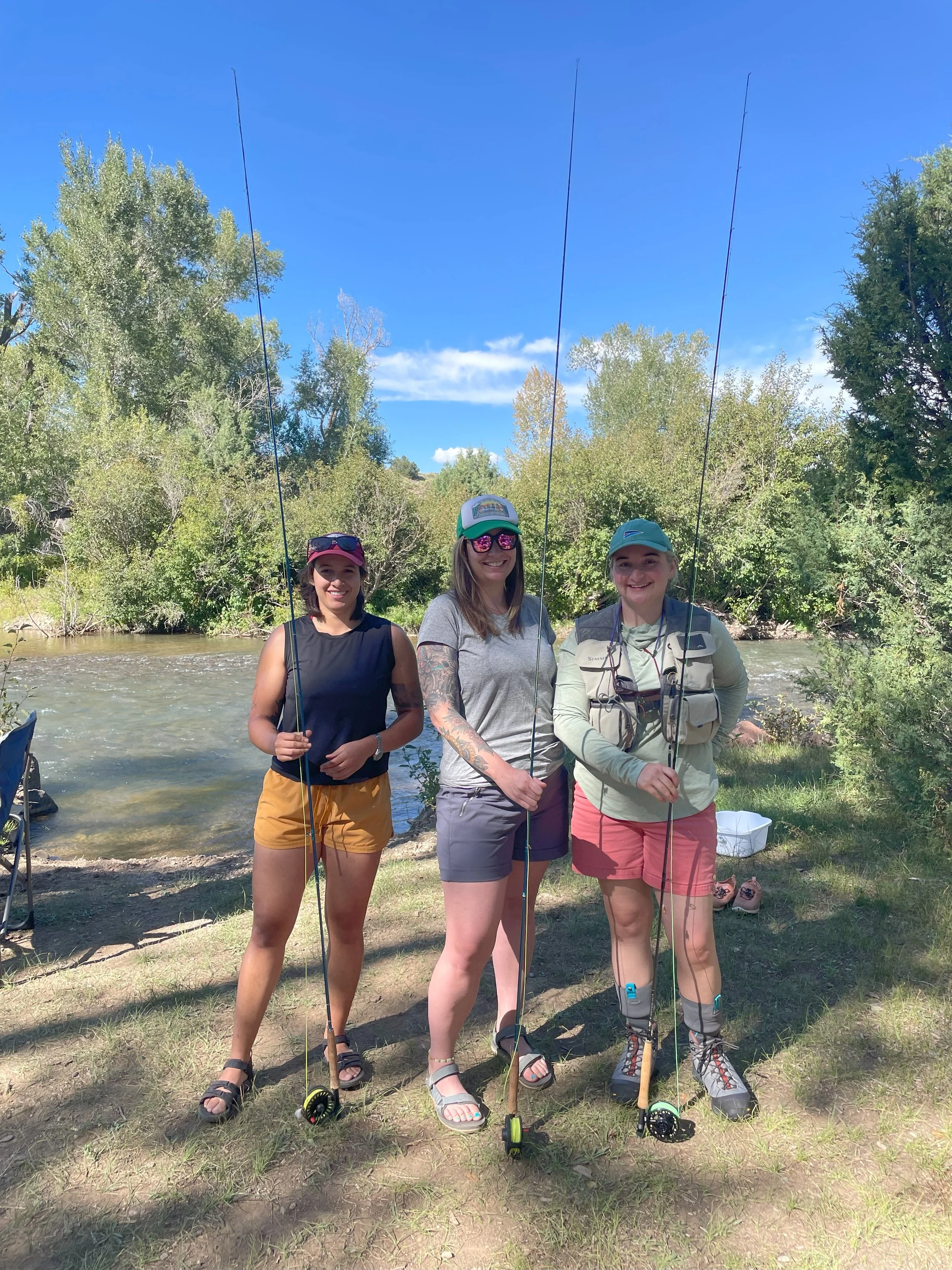 Three women stand on a grassy riverbank holding fishing rods, smiling at the camera, with trees and a river in the background on a sunny day.