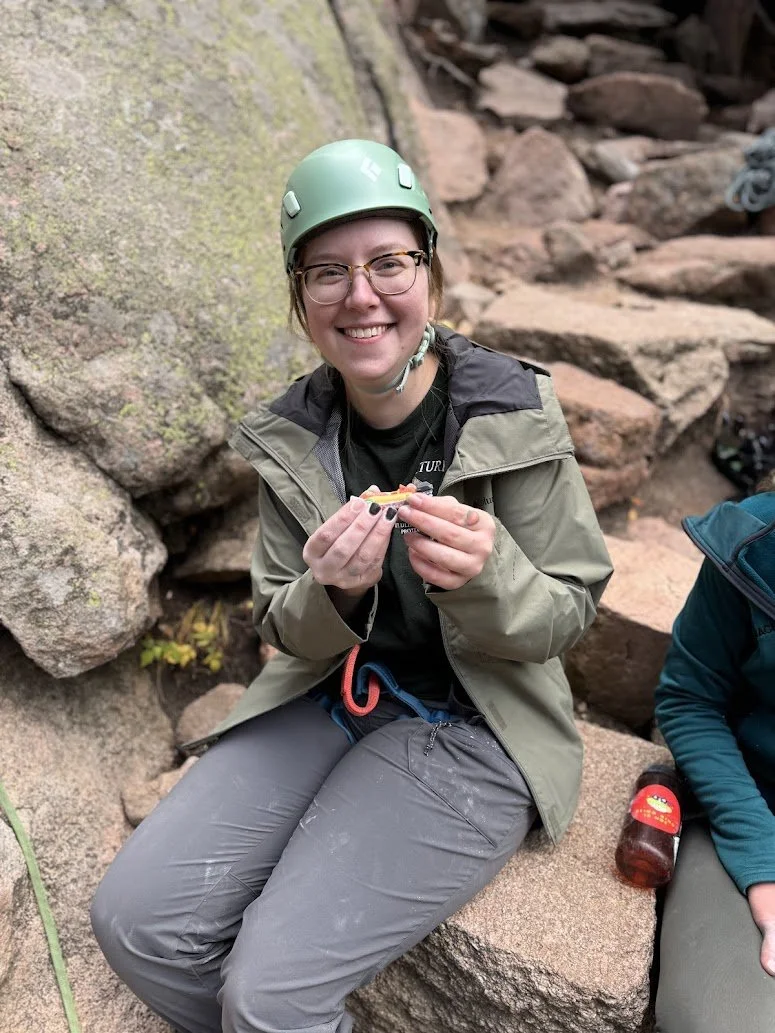 A woman wearing a green climbing helmet, glasses, and outdoor gear sitting on rocks, holding a snack, and smiling at the camera.