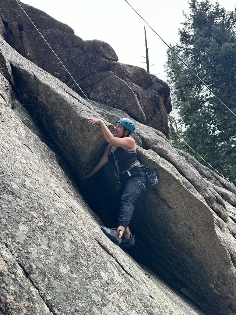 A woman wearing a blue helmet and black climbing gear is rock climbing on a large, sloped granite surface outdoors, with trees and power lines in the background.