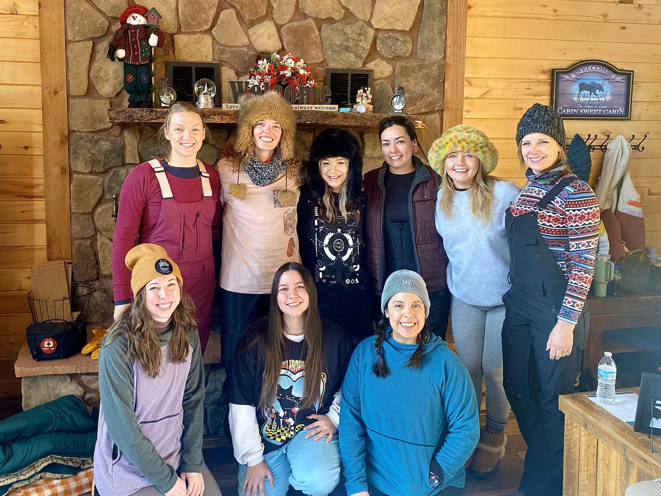 Group of ten women wearing winter clothing, hats, and beanies, smiling indoors in a cozy cabin with wood-paneled walls and a stone fireplace decorated with holiday ornaments.