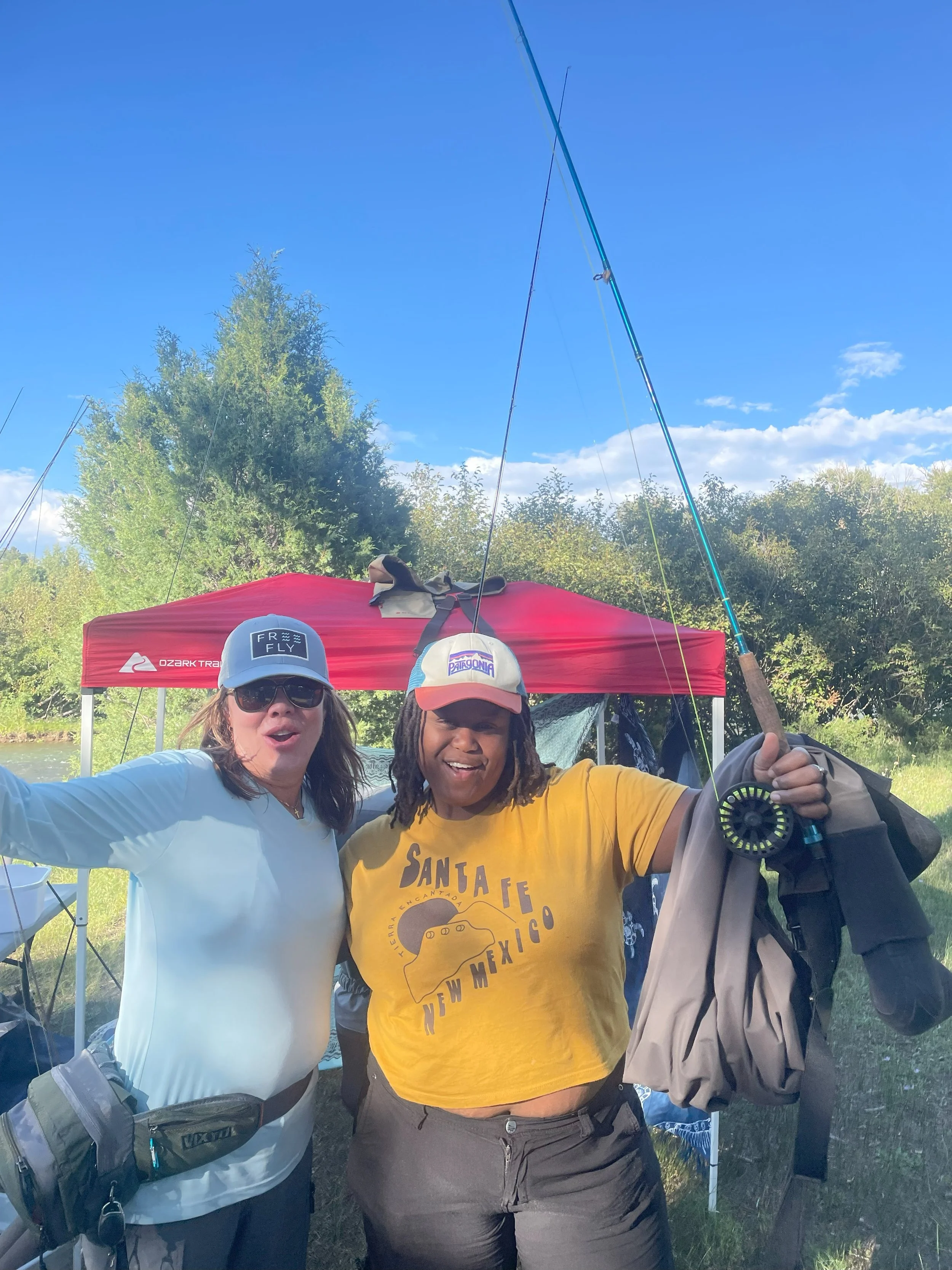 Two women smiling outdoors, dressed for fishing, standing in front of a red canopy with trees and blue sky in the background.