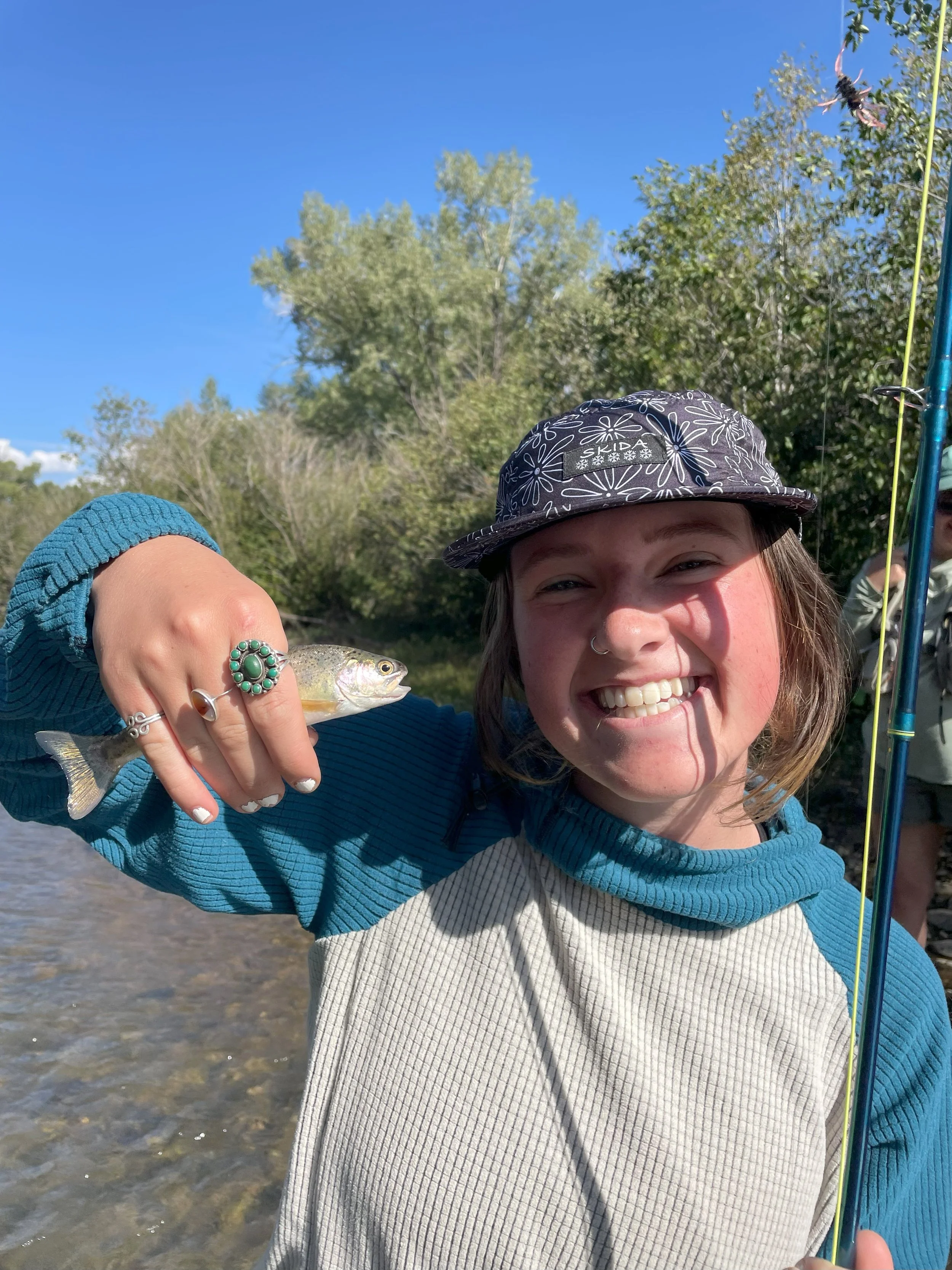 A young woman smiling and holding a small fish she caught outdoors near a river, with trees and a clear blue sky in the background.