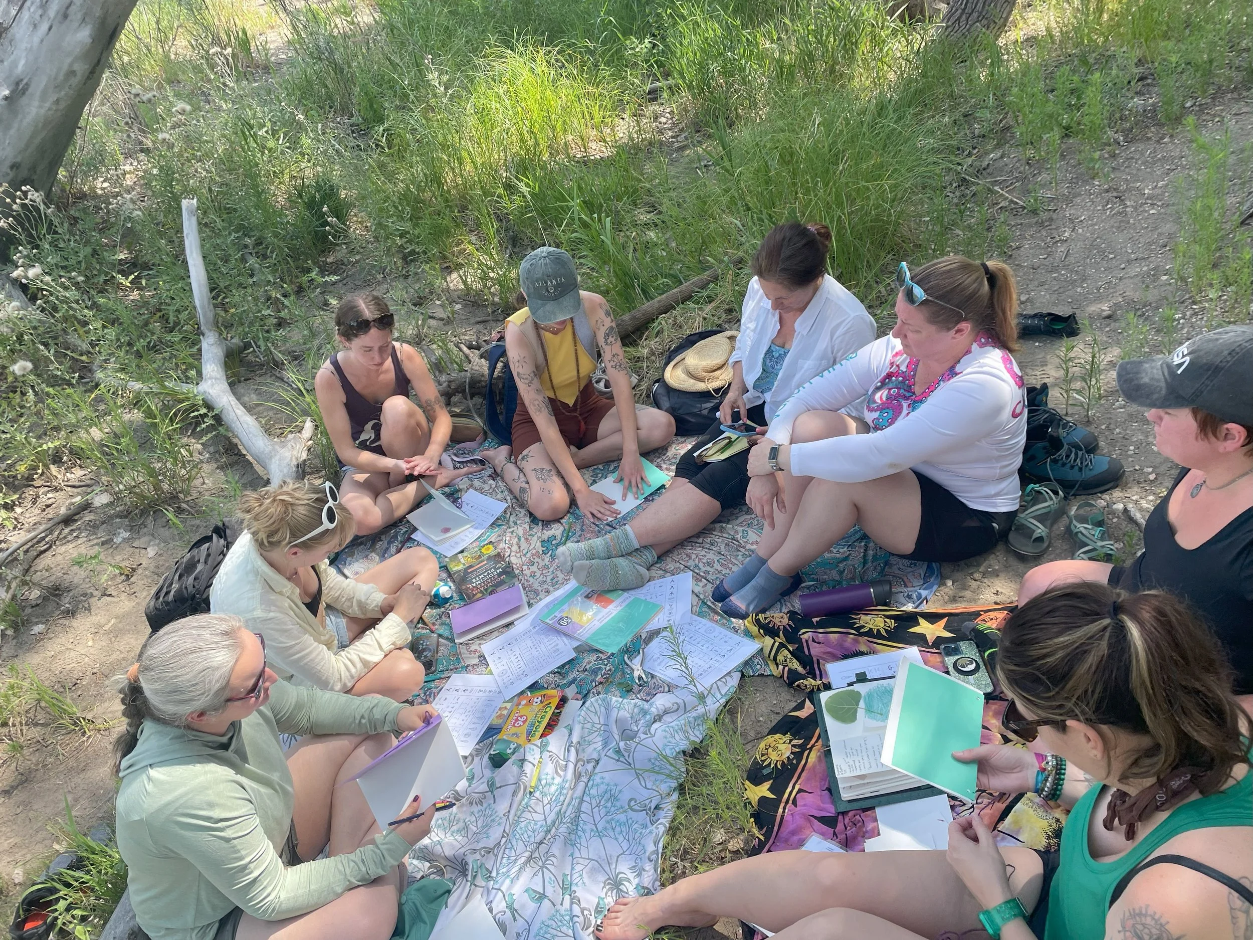 Group of women and girls sitting on a colorful blanket outdoors, surrounded by green grass and trees, with books, notebooks, and art supplies scattered around.