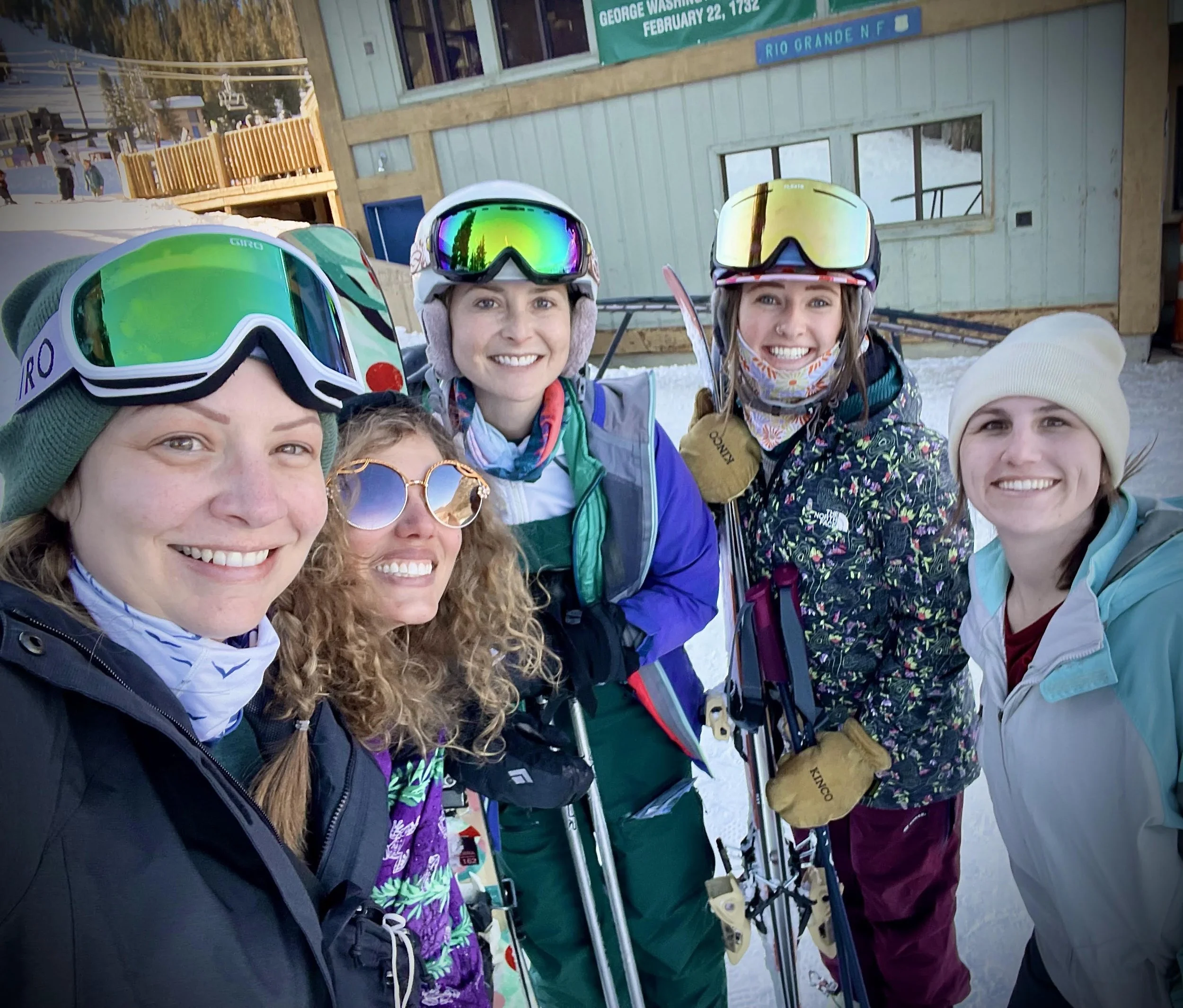 Group of six women in winter ski gear smiling at the camera, standing outdoors near a wooden building with snow on the ground.