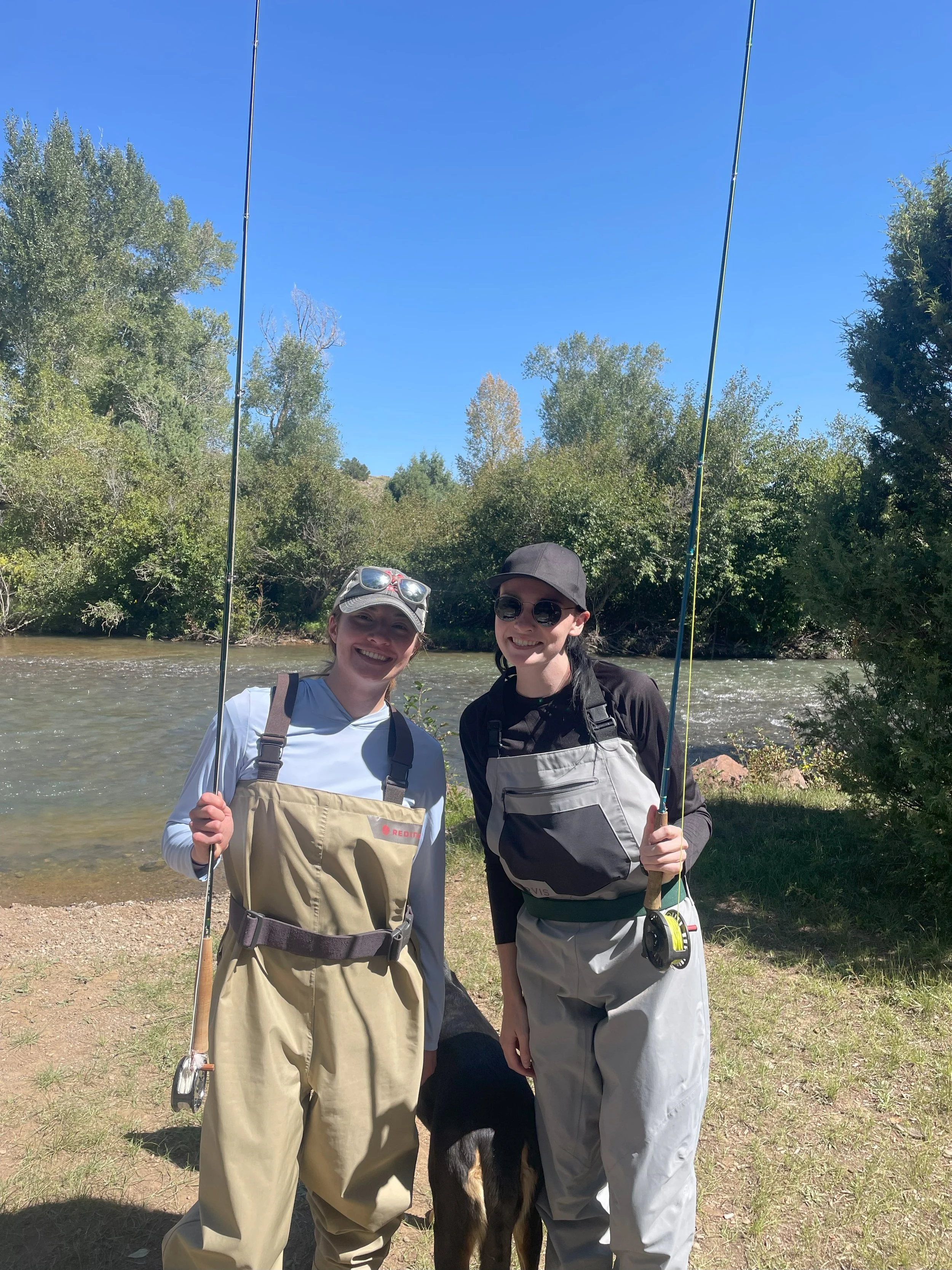 Two women in fishing gear holding fishing rods, standing near a river with green trees and a clear blue sky in the background, smiling at the camera, with a black and brown dog partially visible between them.