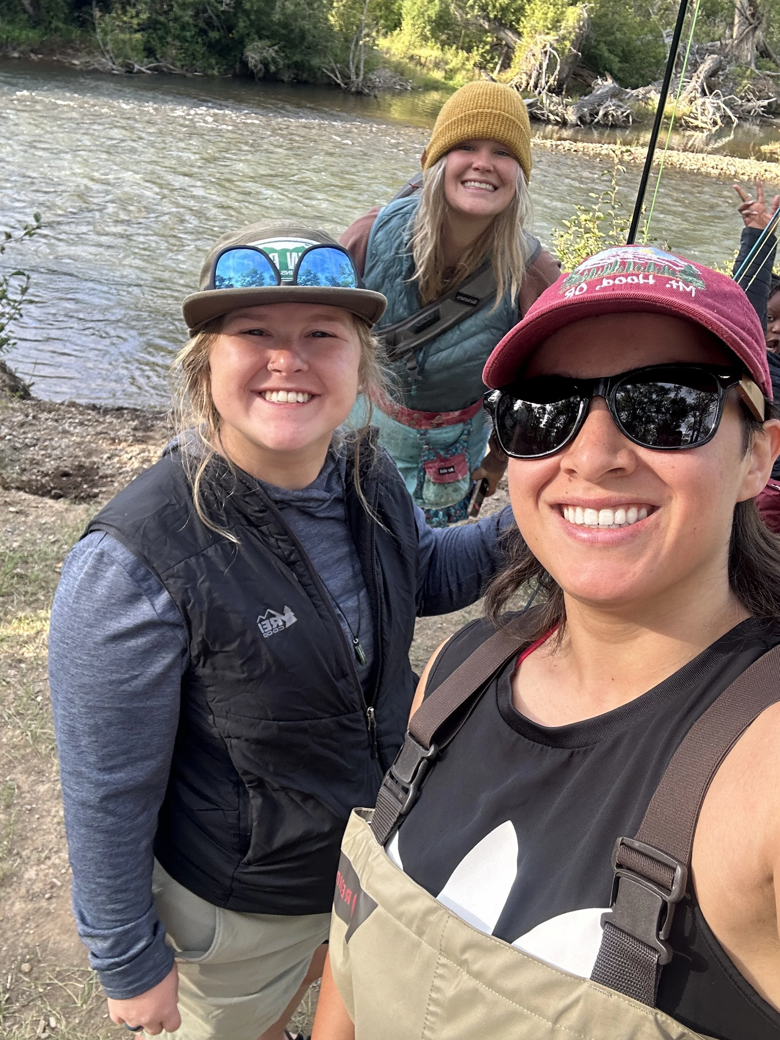 Three women outdoors by a river, smiling and taking a selfie, dressed in casual outdoor clothing and hats.