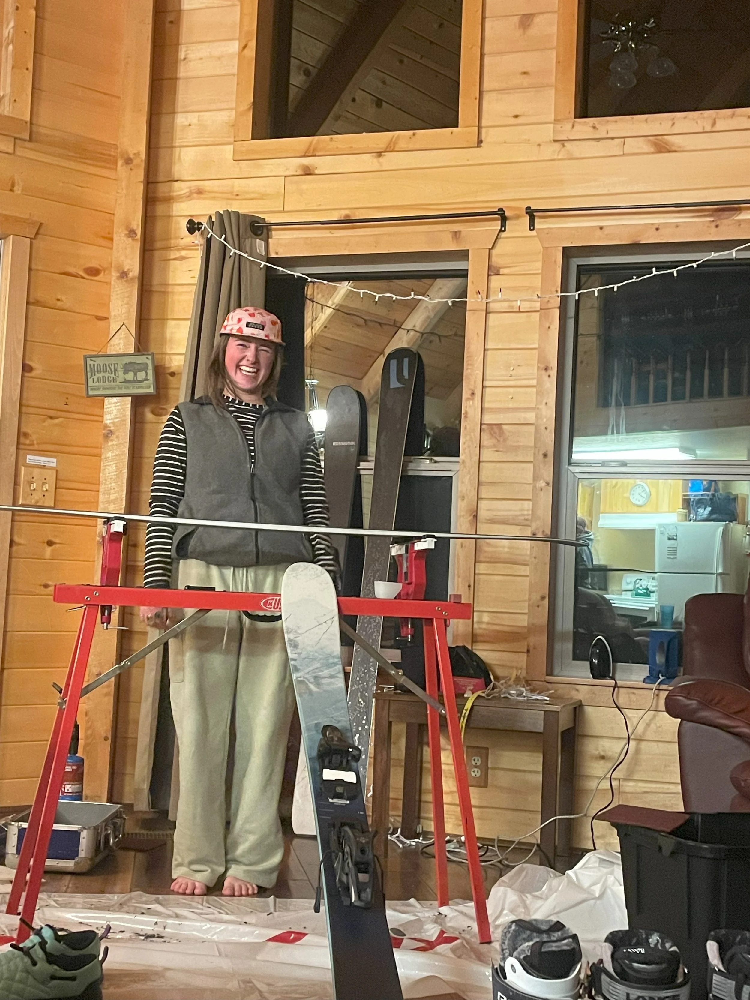 A woman smiling and wearing a helmet, standing indoors in front of ski equipment and a red grooming stand, in a wood-paneled room with string lights and windows.
