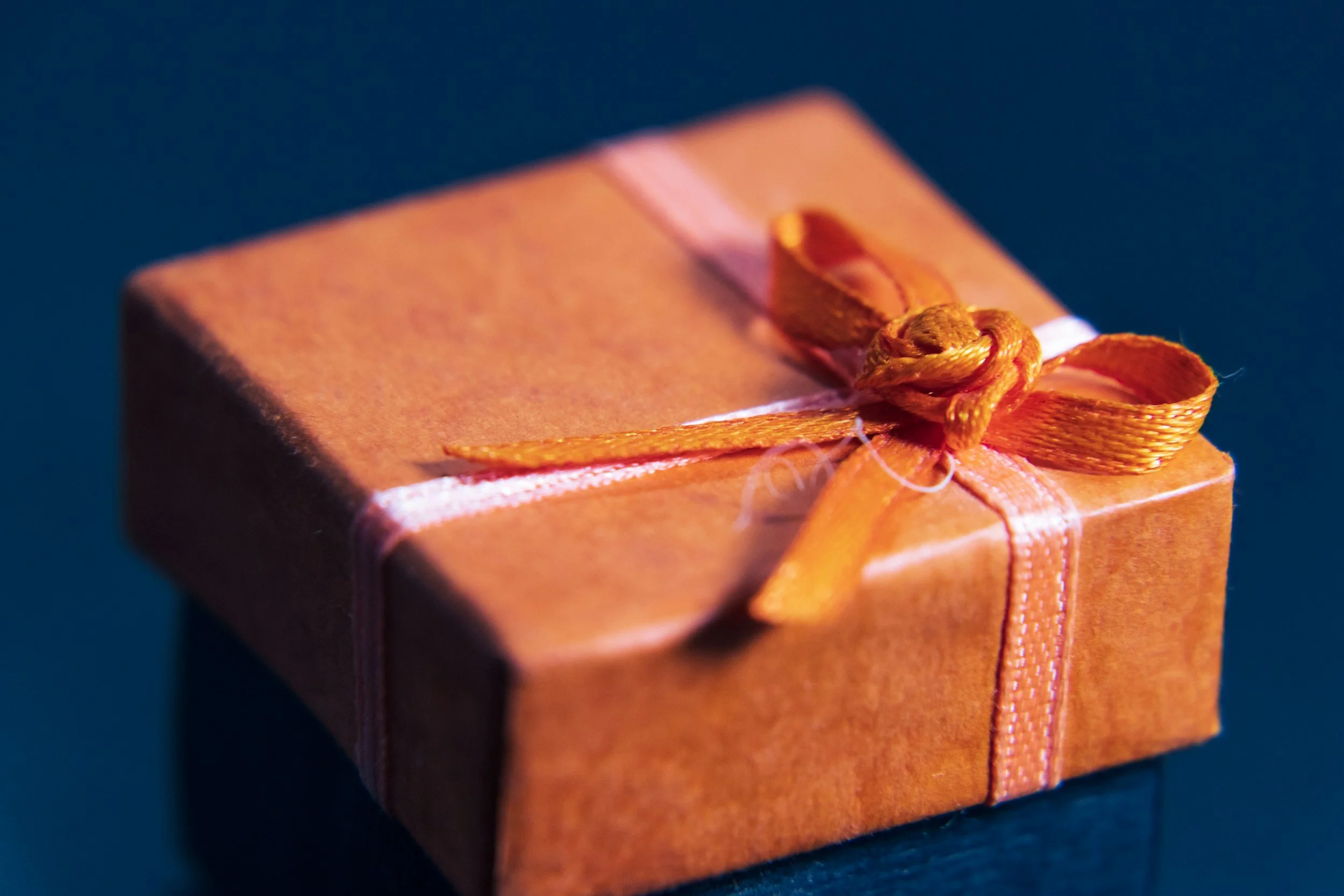 Close-up of a small, square, orange gift box wrapped with an orange ribbon tied into a bow, placed on a dark blue surface.