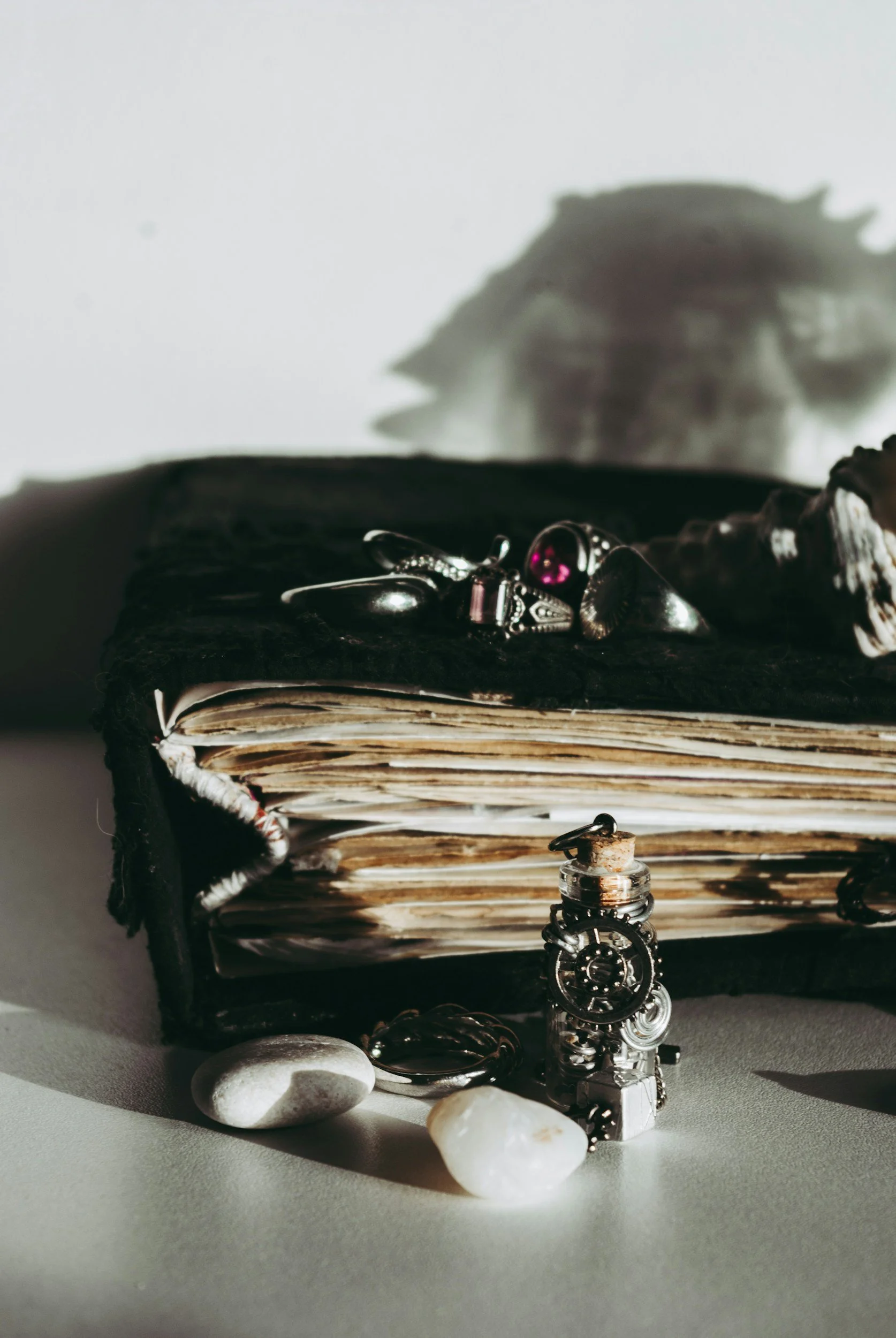 An open vintage book with a black cover and yellowed pages, decorated with rings and jewelry. White stones and a small glass vial with a cork stopper are placed around the book, with jewelry hanging from the stones and the book.