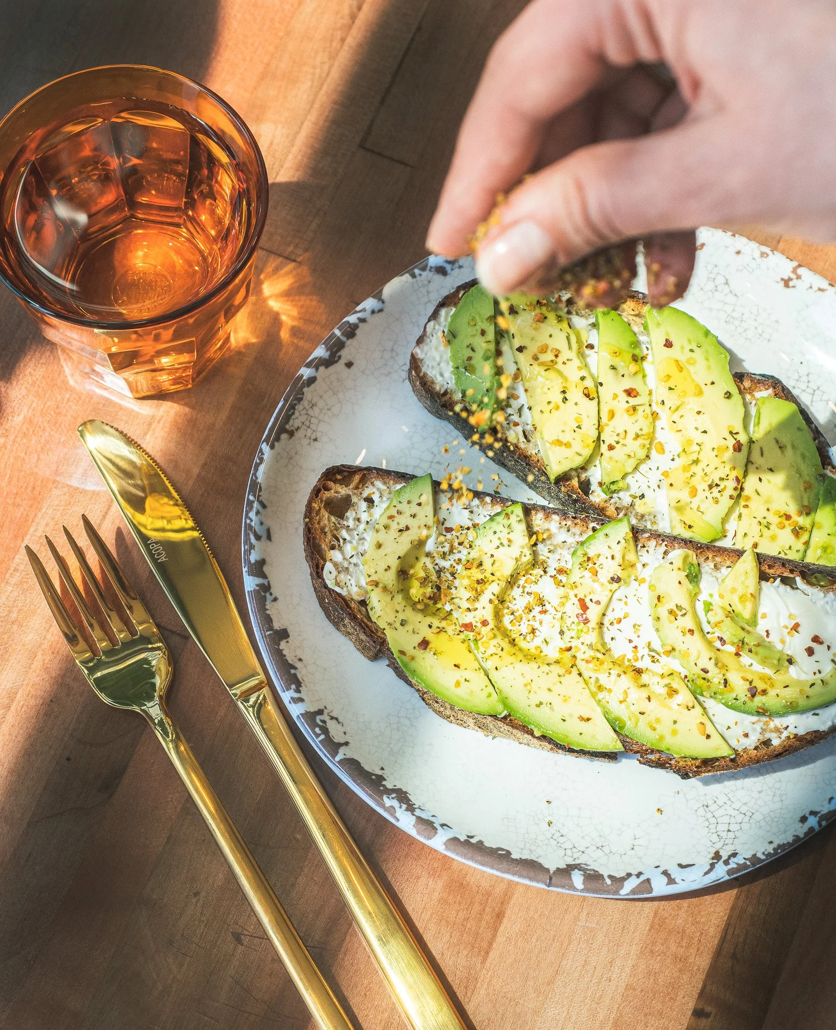 avocado toast on night moves sourdough served on a white plate with gold cutlery and a glass of water. a hand is sprinkling garnish on the toast