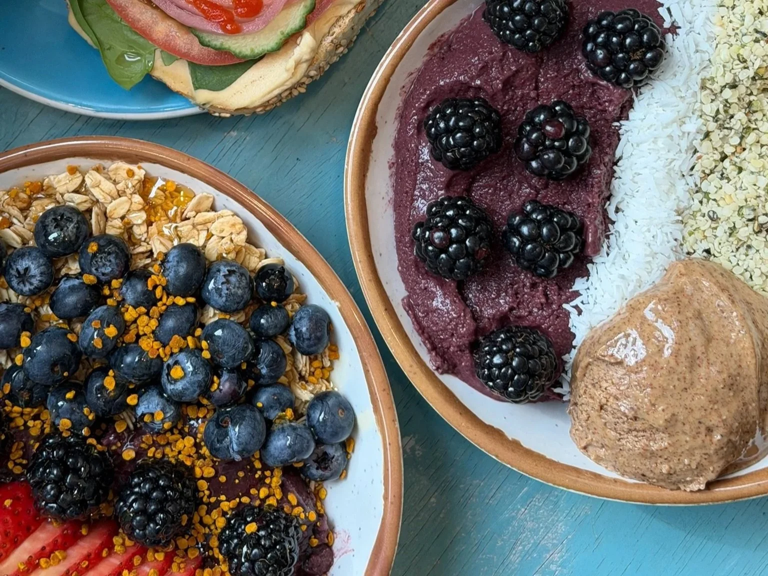 Assorted bom dia acai bowls of colorful fruit, including blueberries, blackberries, coconut flakes, granola, and peanut butter, with a bagel with vegetable toppings on a blue wooden table.