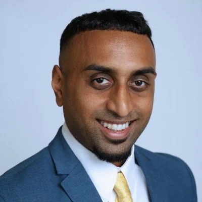 Man wearing a blue suit and yellow tie, smiling against a light background.