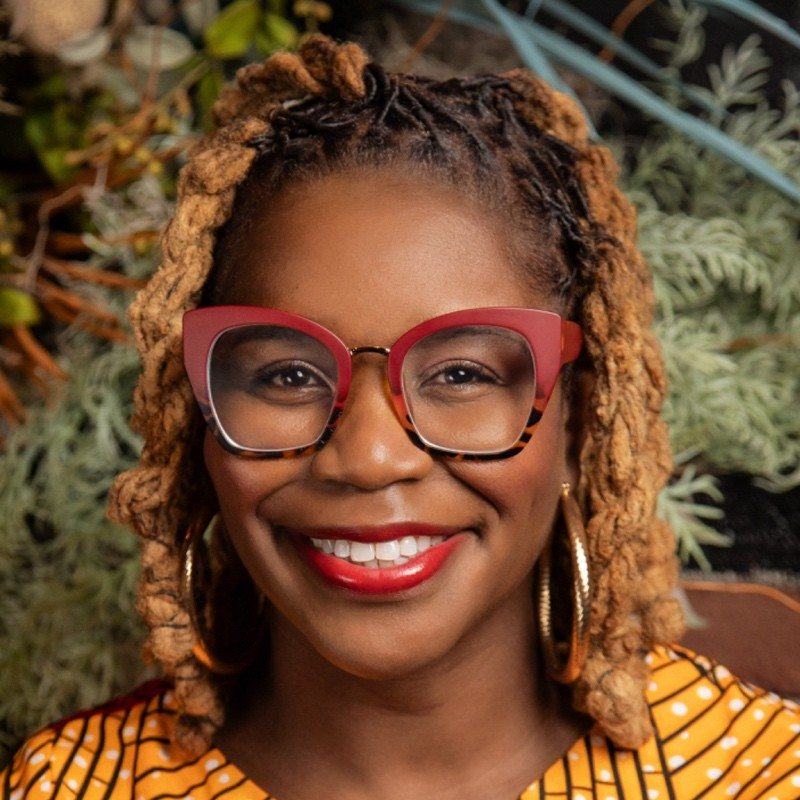 Smiling person with glasses and braided hair in front of greenery background.