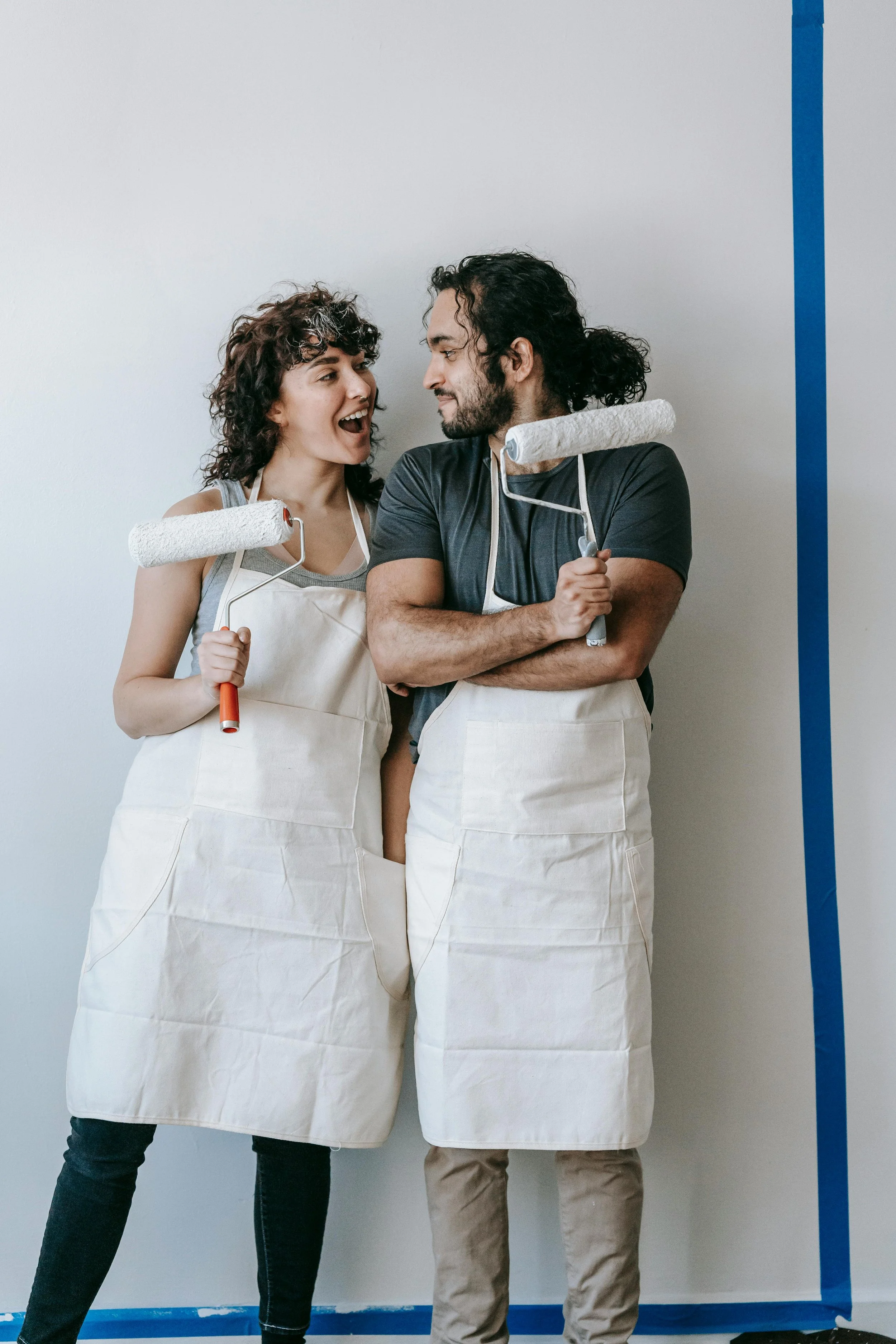 A couple wearing aprons holding paint rollers, standing against a taped-off wall.