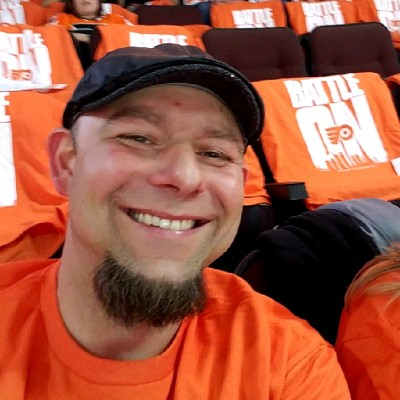 Man smiling, wearing an orange T-shirt and a black cap, seated in an arena with "Battle On" text on the seats.