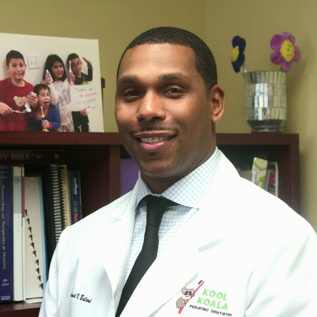 A man in a white lab coat with "Kool Koala Pediatric Dentistry" logo, standing in an office with books and a vase of flower decor.