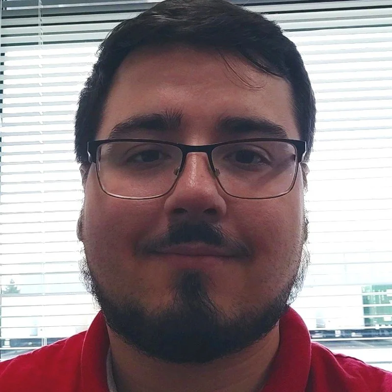 Close-up of a man with glasses, dark hair, a beard, and a mustache, wearing a red shirt in front of window blinds.