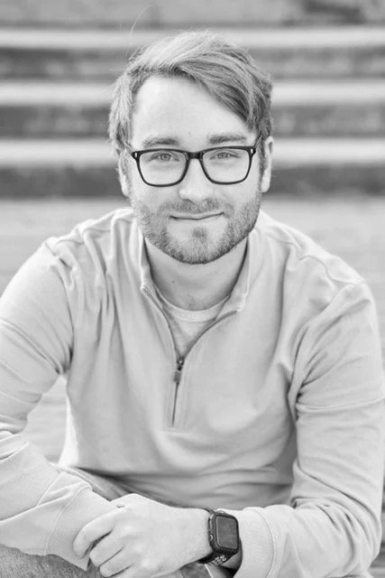 A young man with glasses and a beard smiling, sitting outdoors in front of steps, wearing a light-colored zip-up jacket and a smartwatch.