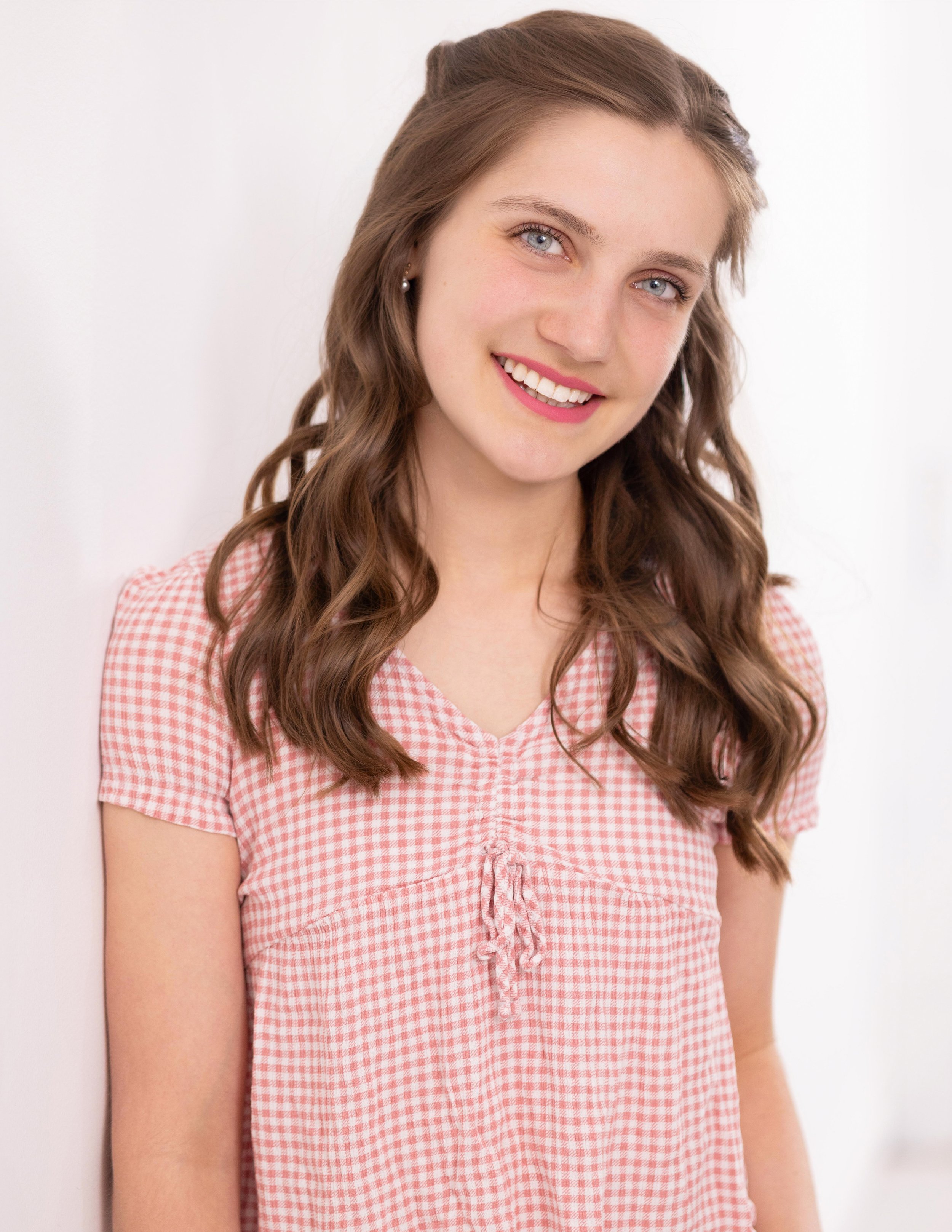 Portrait of a young woman with long, wavy brown hair wearing a pink and white checkered shirt, smiling against a plain white background.