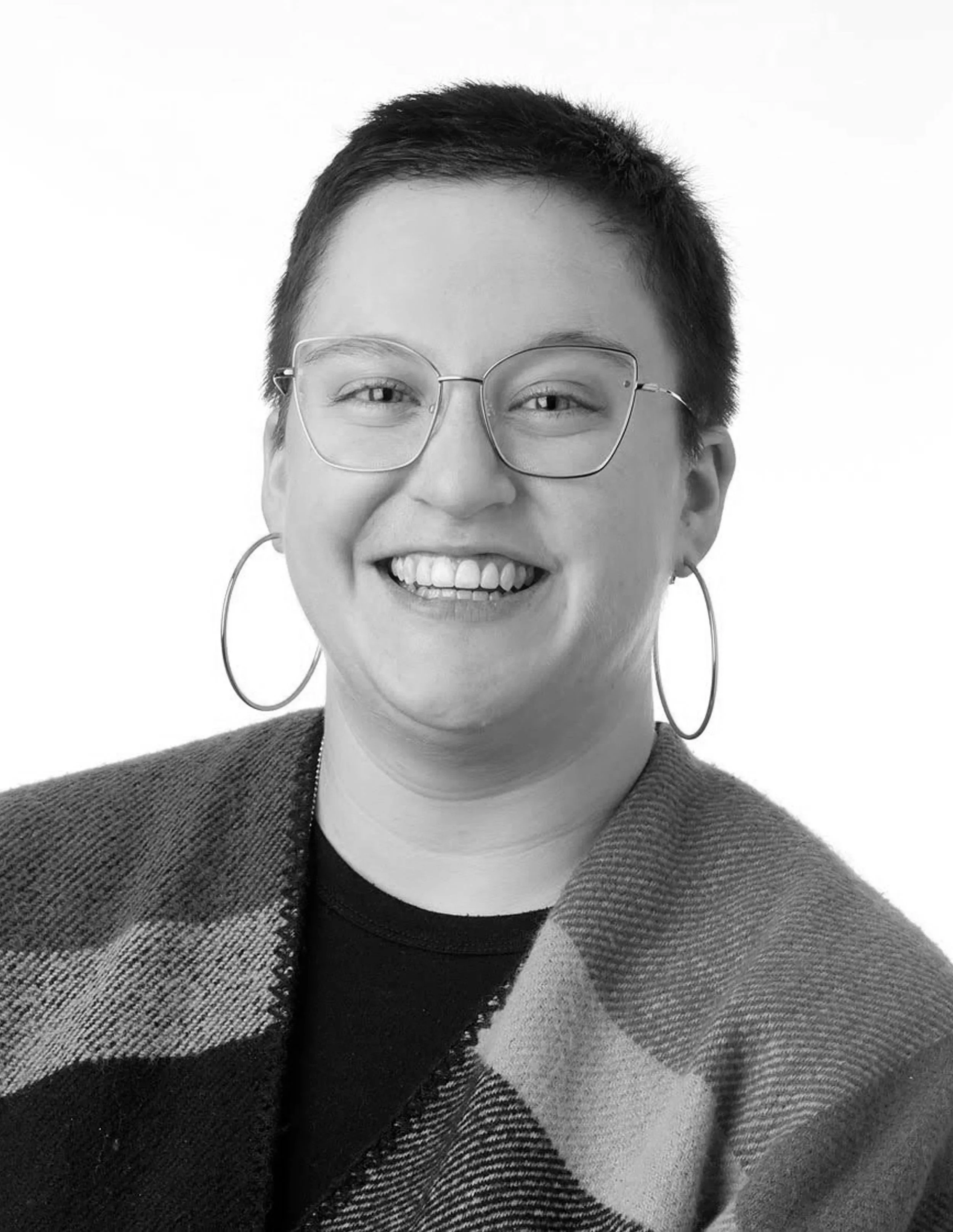A smiling person with short dark hair, wearing large round glasses, hoop earrings, a black shirt, and a dark patterned coat, against a white background.