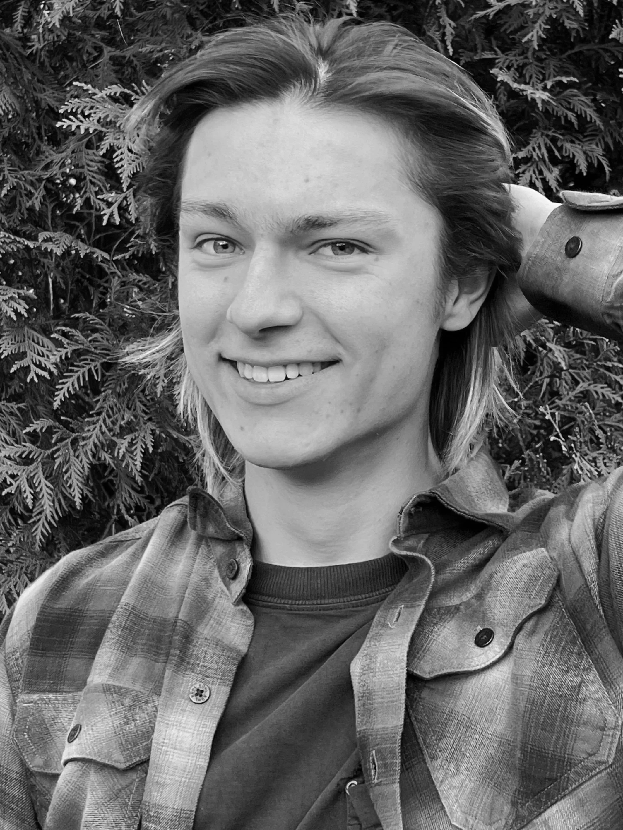 A close-up black and white photo of a young person with shoulder-length hair, smiling, wearing a plaid shirt layered over a dark t-shirt, with trees and foliage in the background.