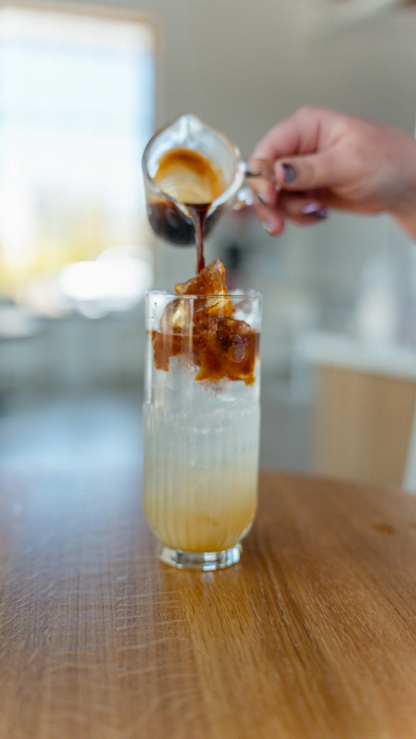 A hand pouring coffee into a tall glass with ice and a slice of lemon, on a wooden table.