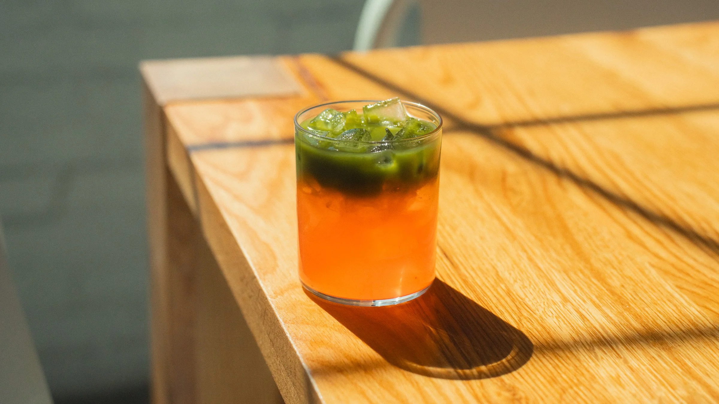 A glass of iced tea with ice cubes and green liquid on top, placed on a wooden table.