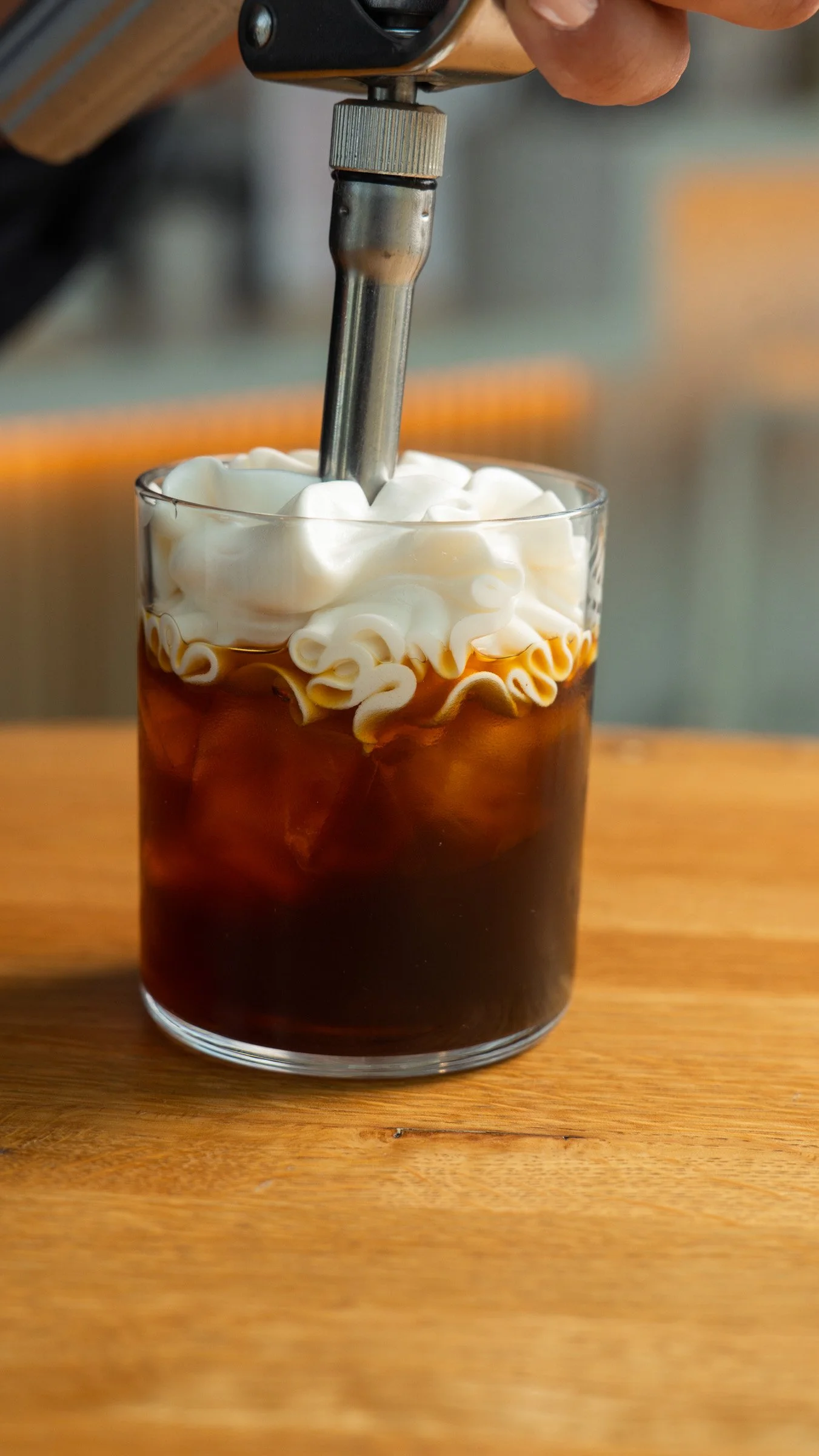Close-up of a glass of iced coffee topped with whipped cream, with a metal spoon stirring the drink, on a wooden surface. Award winning coffee shop