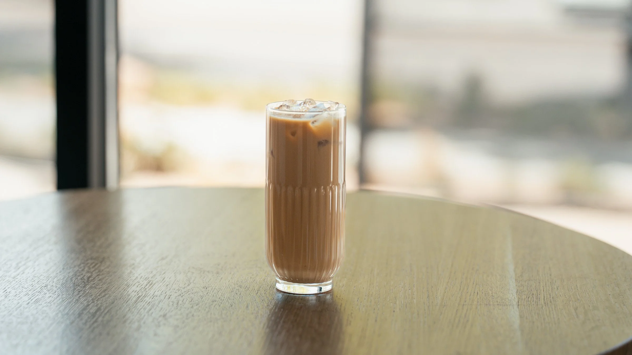 A tall glass of iced chai latte on a round wooden table near a window, with a blurry outdoor background.