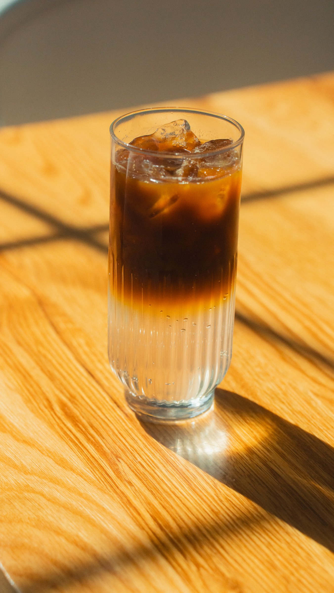 Glass of iced coffee on a wooden table with sunlight and shadows.
