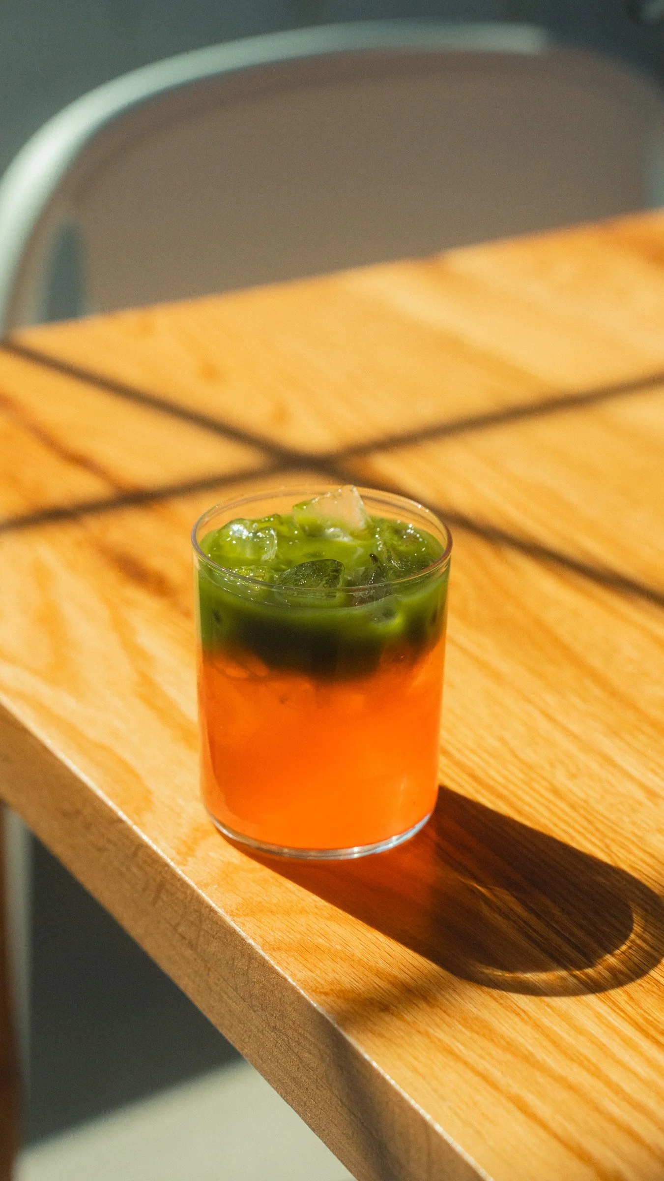 A glass of tea with ice and green syrup on top, placed on a wooden table with a shadow cast to the right.