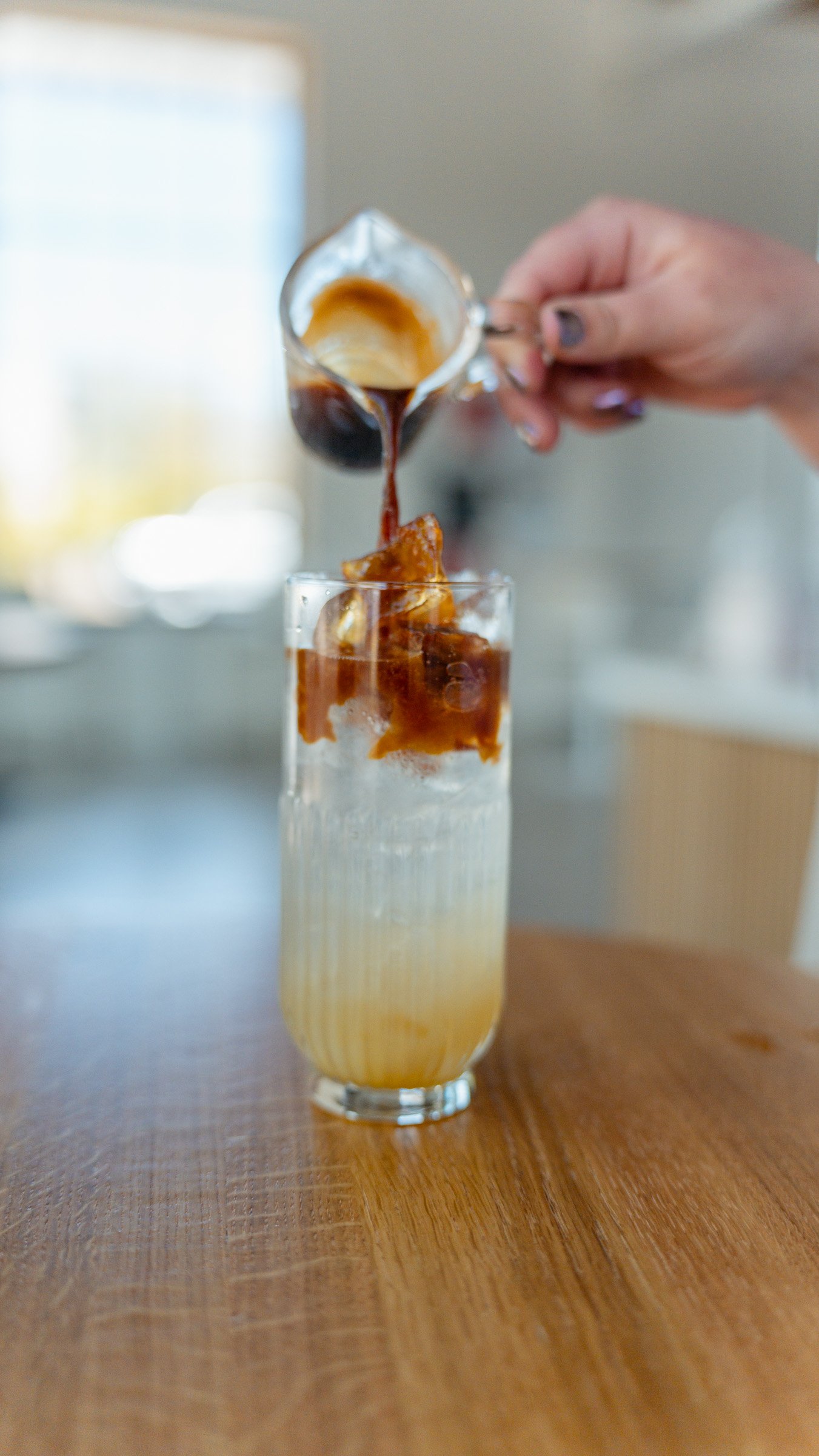 Person pouring coffee into a tall glass with ice, on a wooden table.