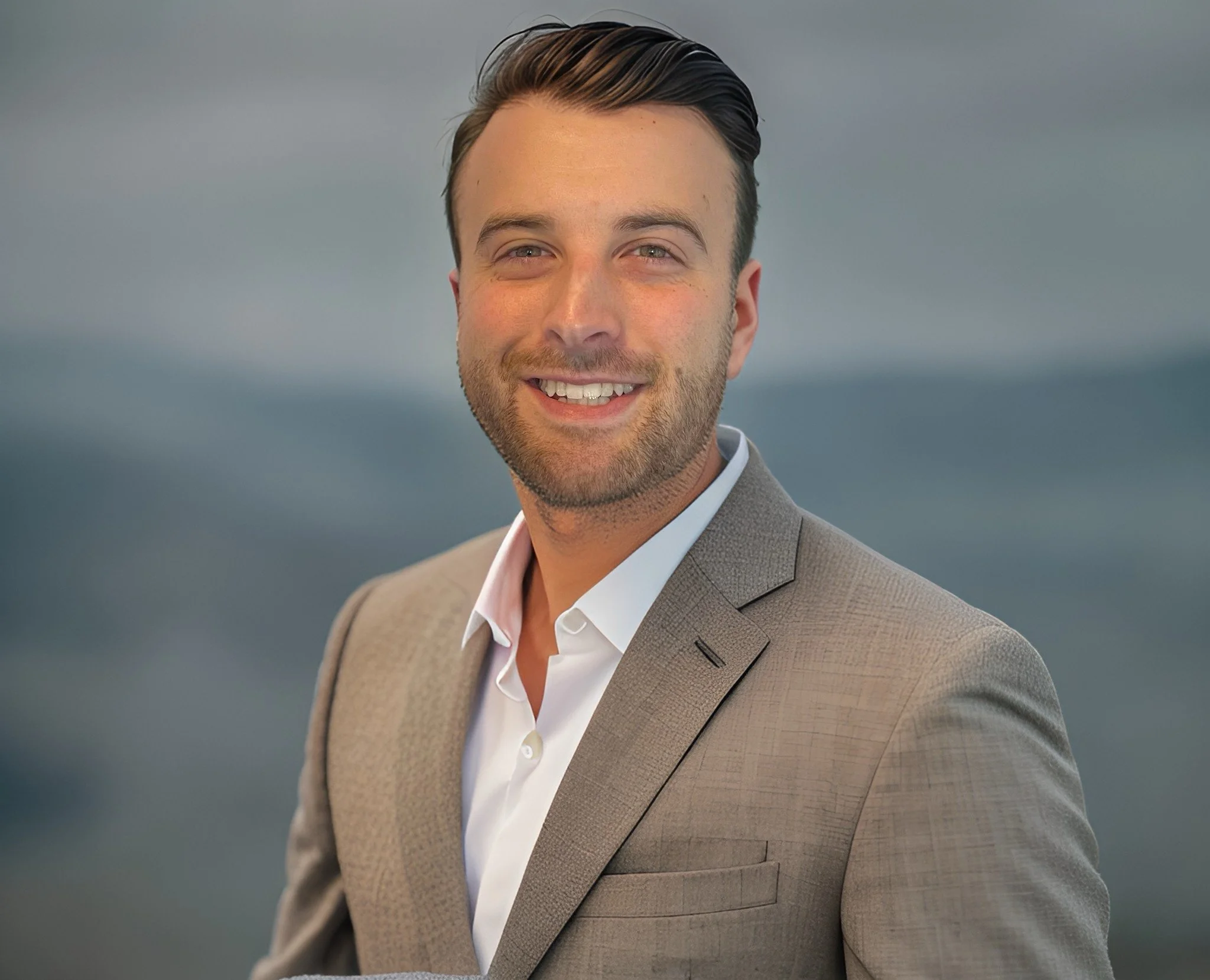 A smiling man in a beige suit with a white shirt, standing outdoors with a blurry mountain landscape in the background.