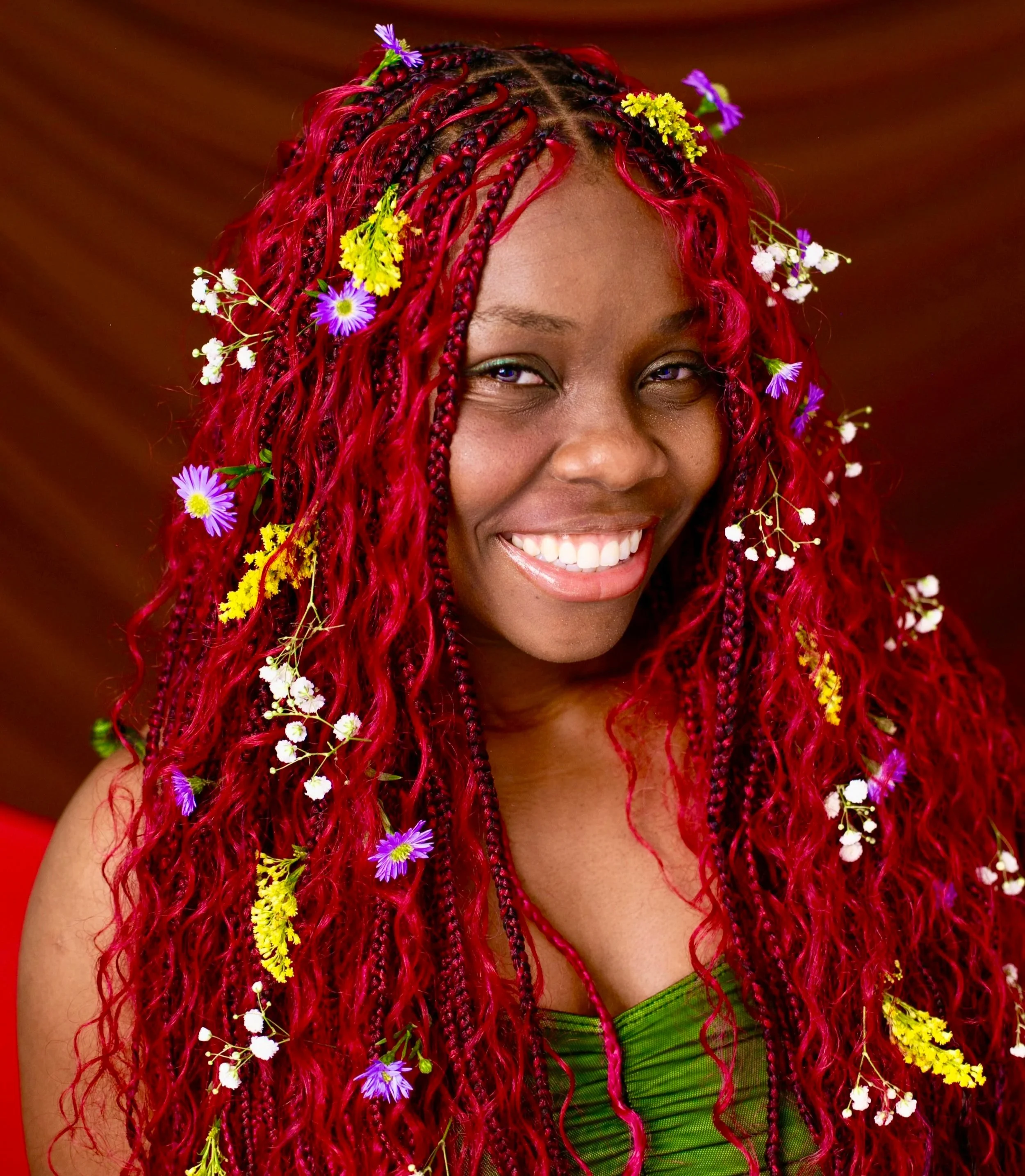A portrait picture of Magdalene smiling. They’re a dark-skinned Black femme in a green top, wearing red long boho braids decorated with flowers of different colors.