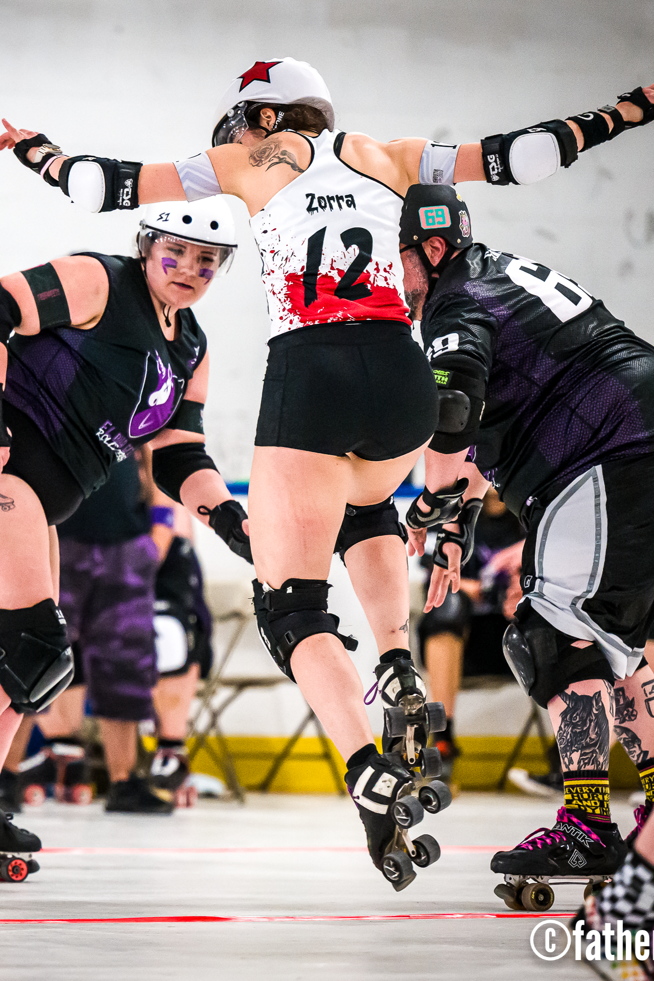 Roller derby players in action, with one wearing a white helmet and a jersey numbered 12, and others around her on roller skates.
