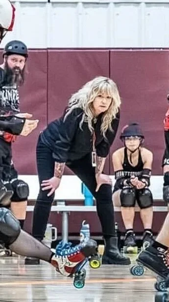 A group of people roller skating indoors, with one woman bending down in the center, and children in the background wearing helmets and protective gear.