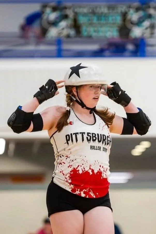 A female roller derby player adjusts her helmet inside a roller skating rink. She wears a white tank top with "PITTSBURGH ROLLER DERBY" written on it, black shorts, and protective gear including wrist guards and elbow pads. Her tank top has red splatter design.