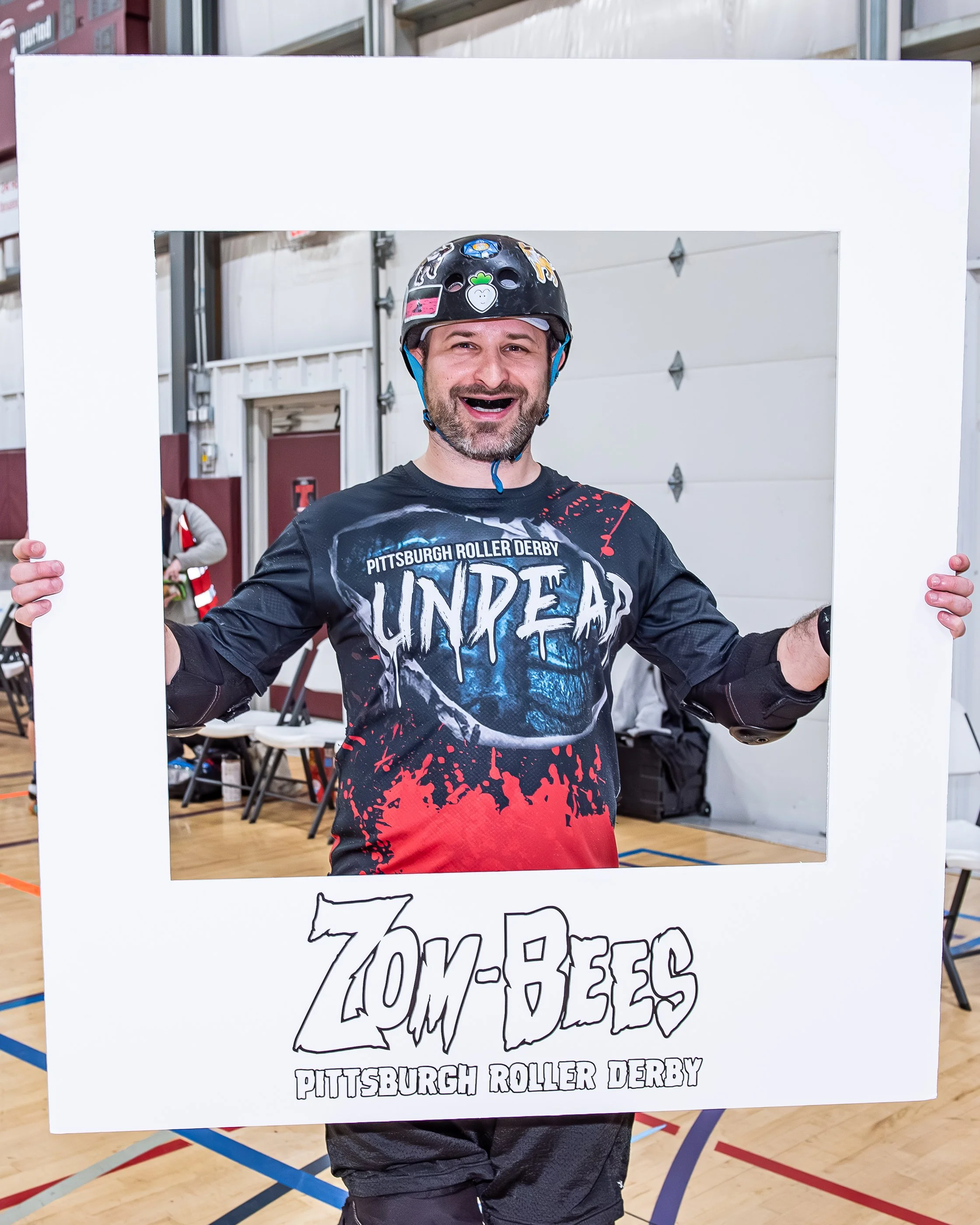 A man wearing a helmet and a Pittsburgh Roller Derby Zombees shirt, holding a large white photo frame with 'Zombees Pittsburgh Roller Derby' written on it, smiling at an indoor roller derby event.